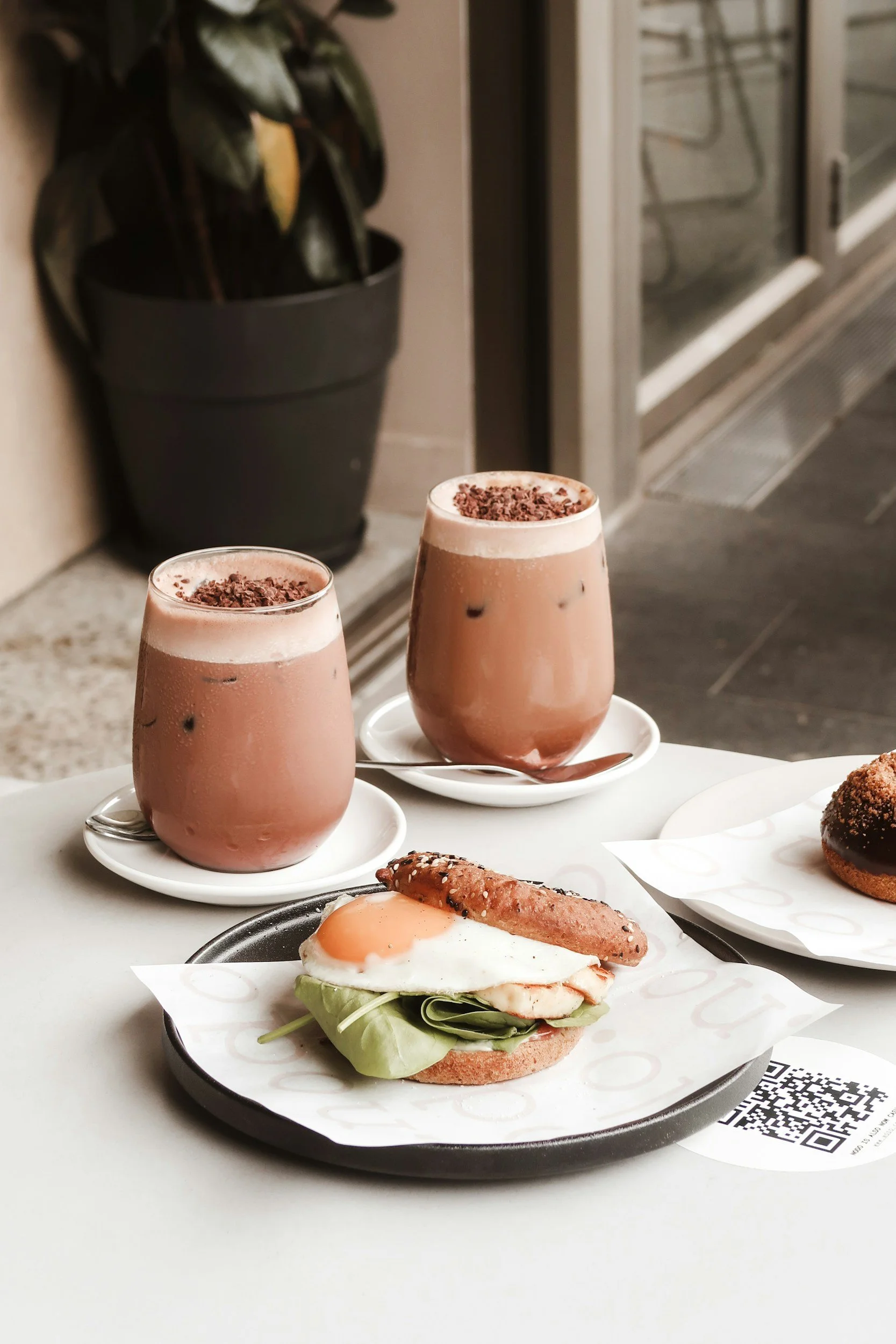 Two glasses of chocolate smoothies topped with chocolate shavings on white saucers, a breakfast plate with a fried egg, a sausage link, next to a breakfast sandwich with lettuce, tomato, egg, and bun, and a croissant on a dark tray.