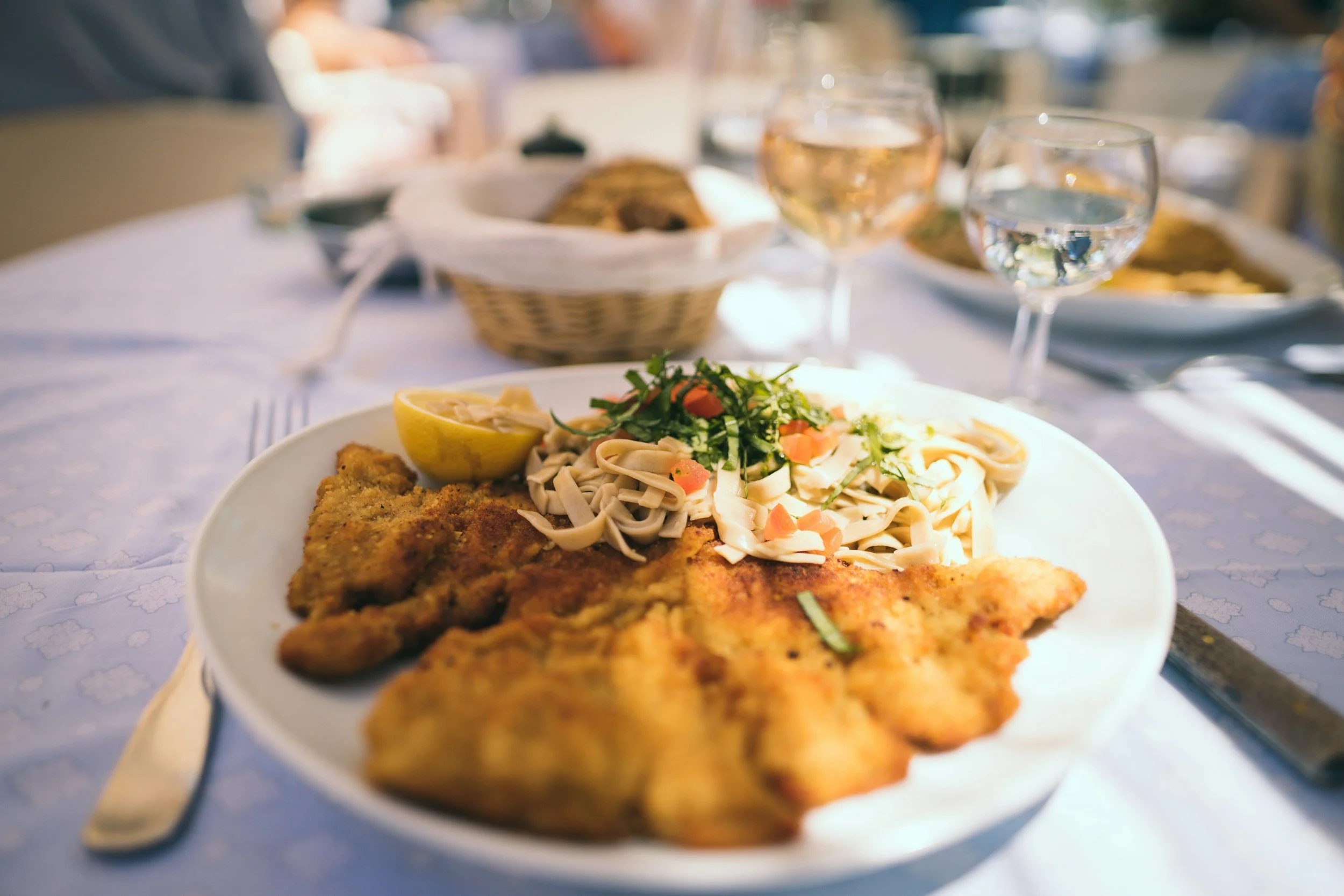 Close-up of a plate with breaded fish fillet, lemon wedge, and pasta salad garnished with herbs, on a table set for a meal