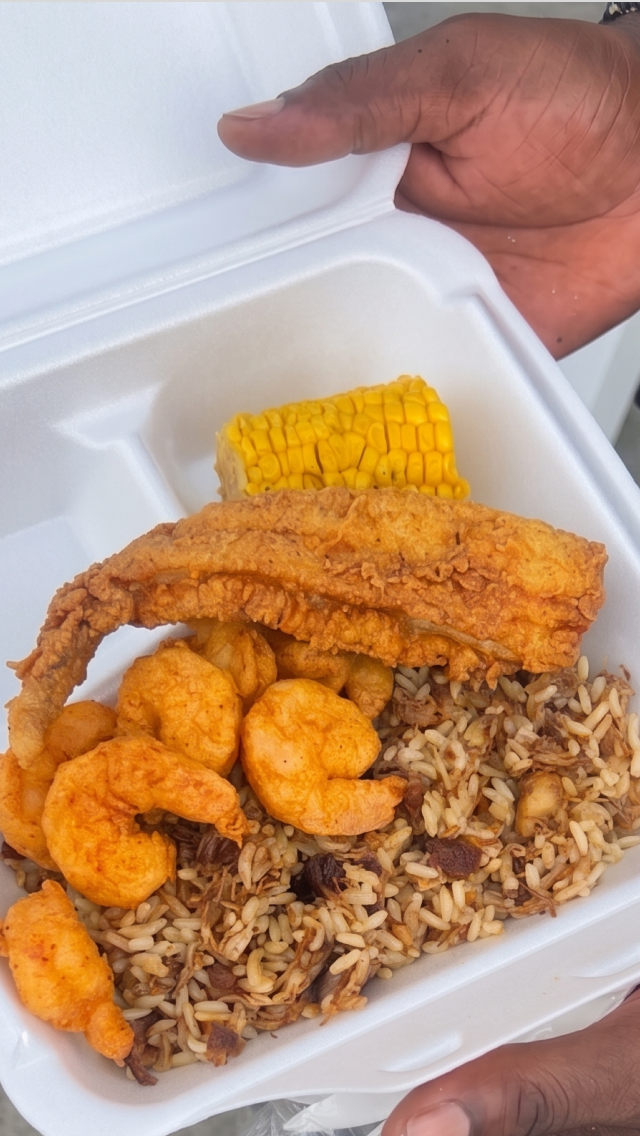Fried shrimp, a piece of fried fish, corn on the cob, and rice with beans in a white foam container.