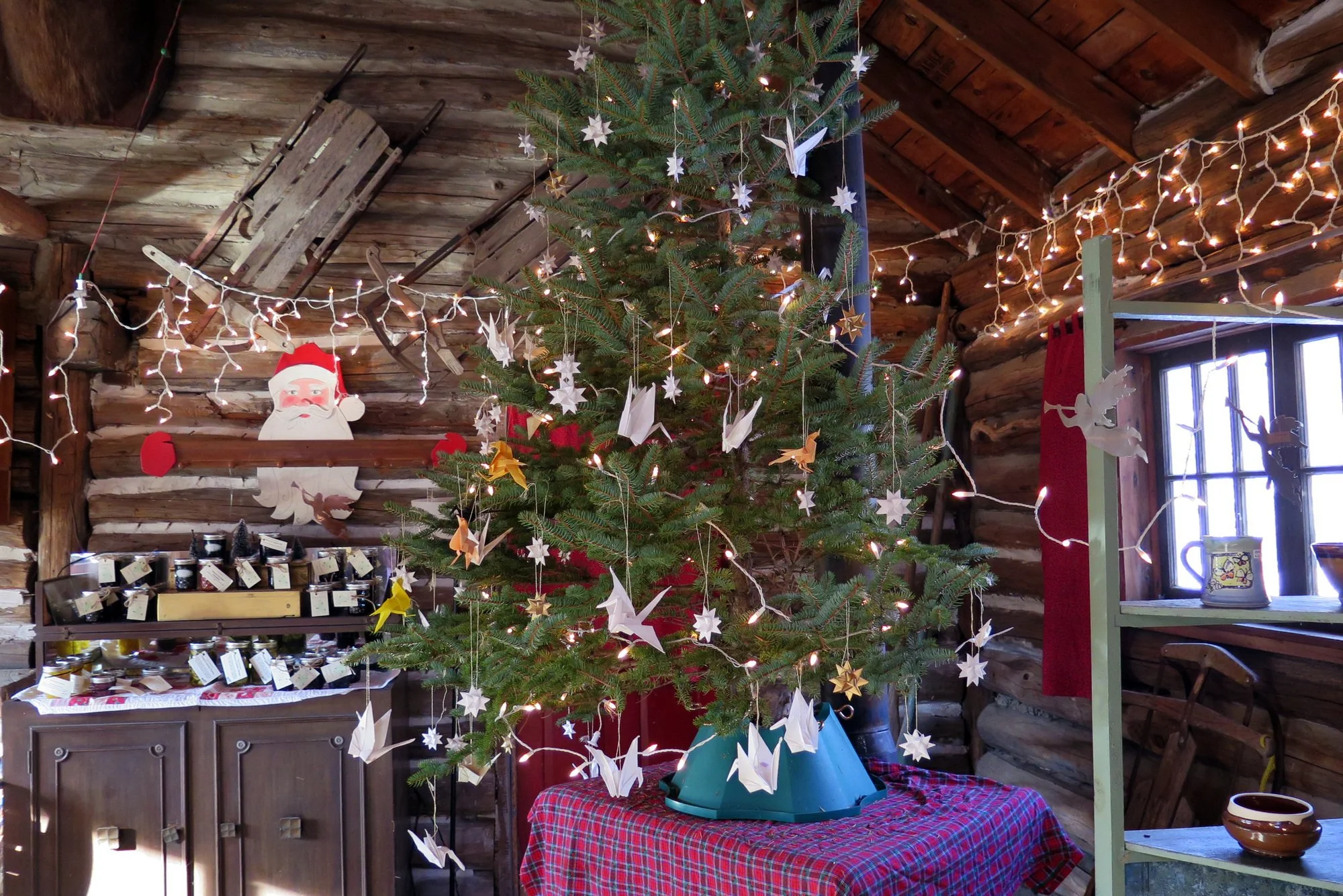 A decorated Christmas tree with white paper stars and string lights inside a log cabin, with a Santa decoration on a wall, a window with snow outside, and a table with jars and other items.