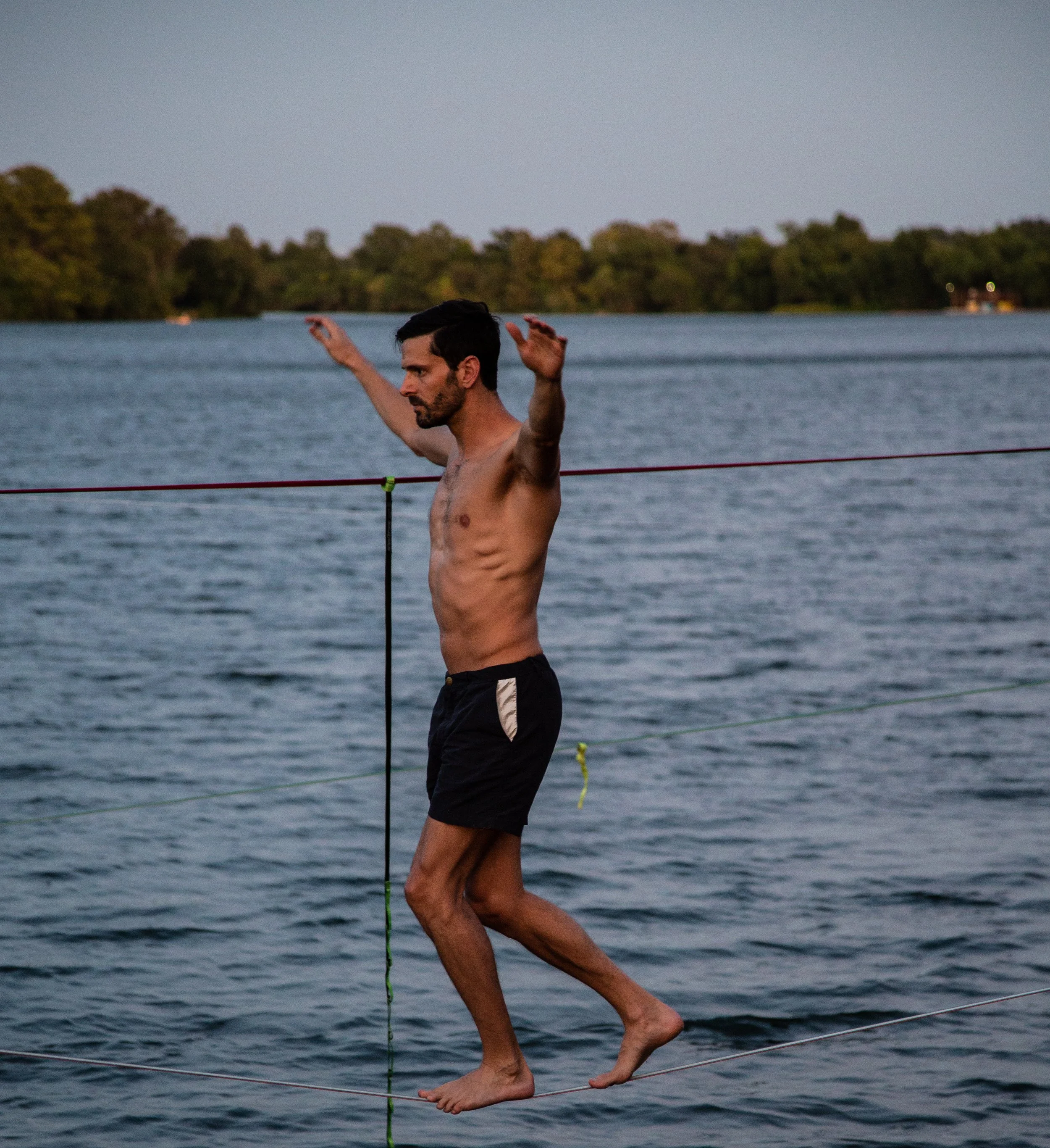 A man walking on a slackline over a body of water at sunset, balancing with arms outstretched.