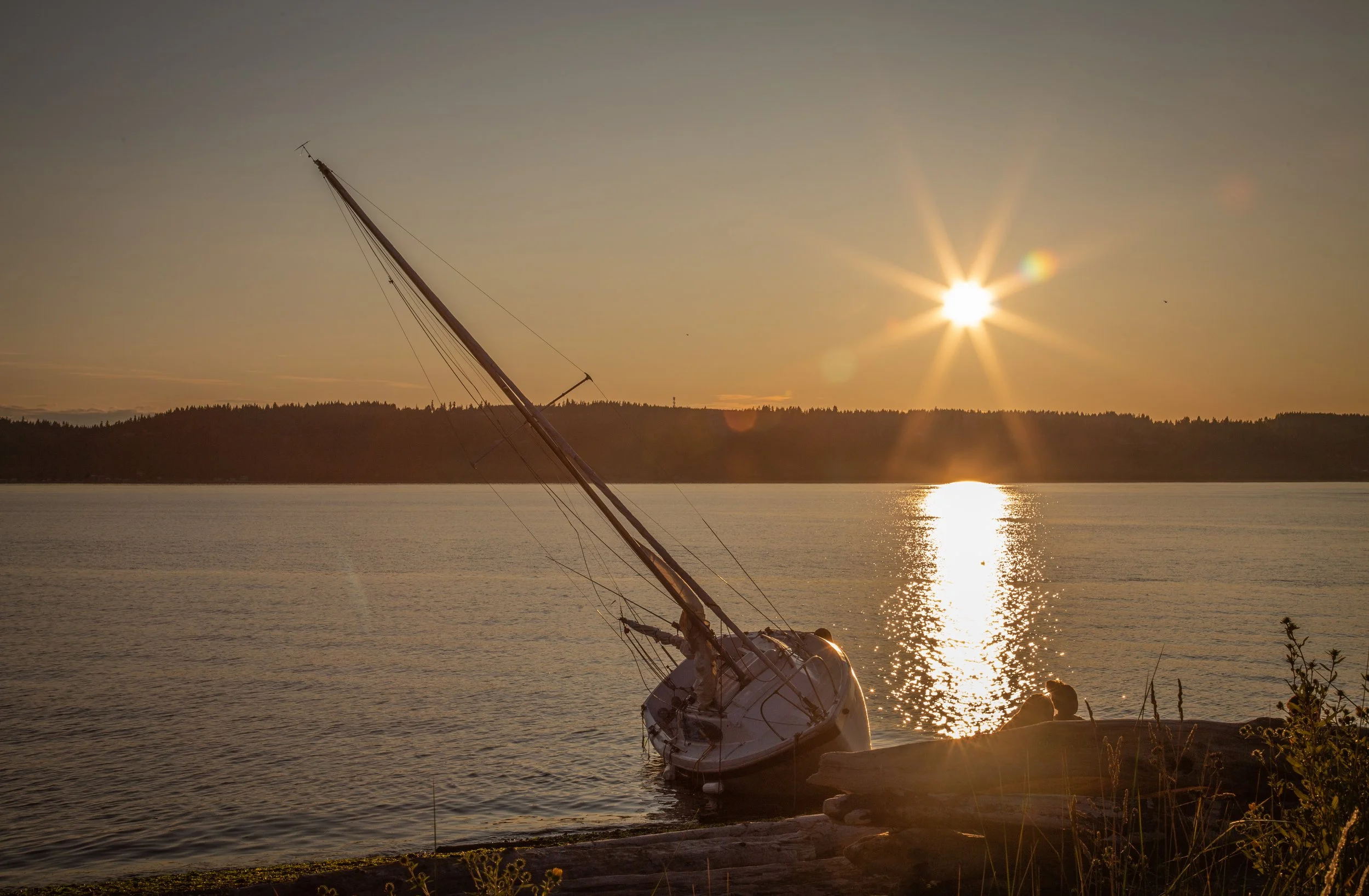 A sailboat tilted on its side near the water's edge during sunset, with the sun reflecting on the calm lake, and a forested shoreline in the background.