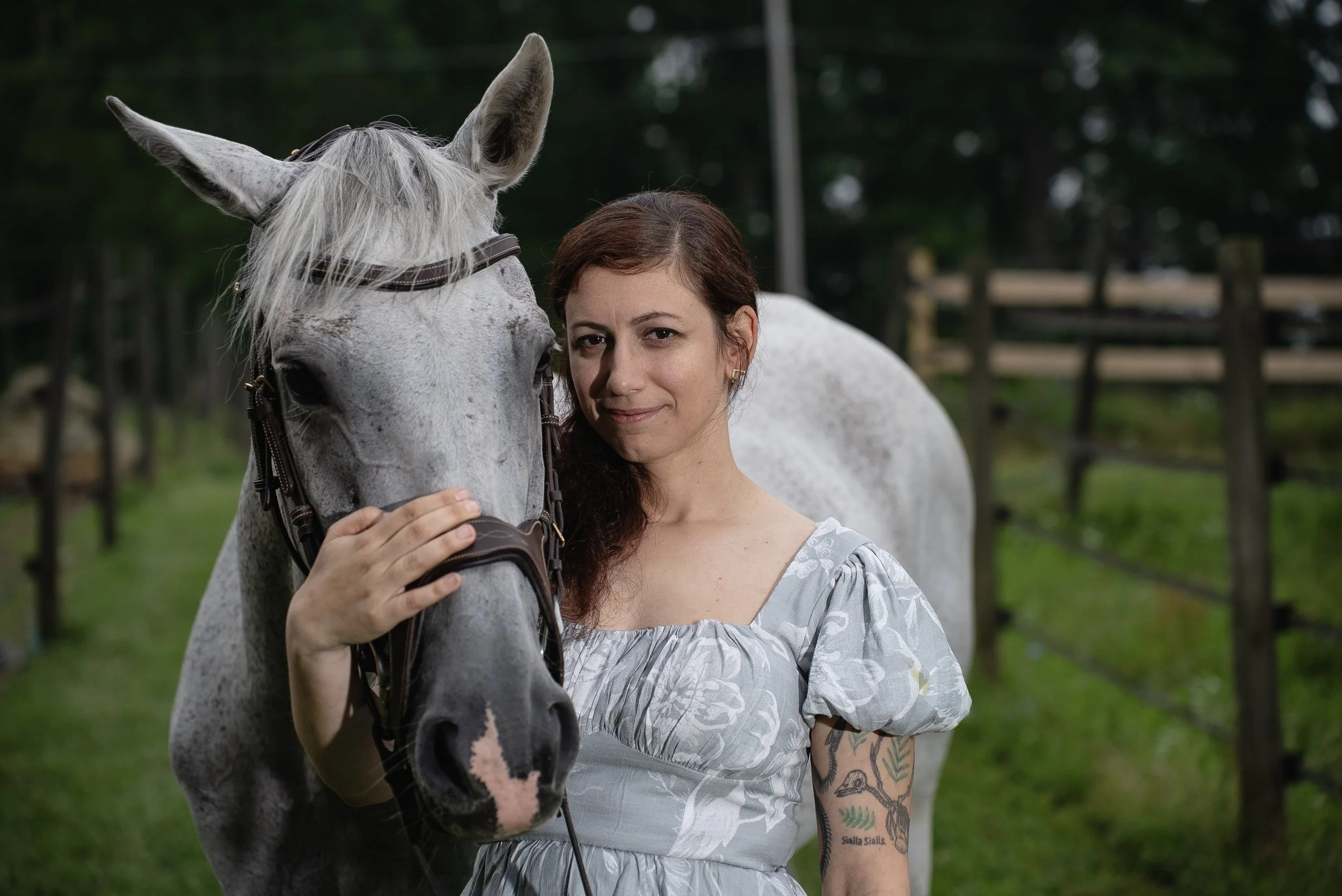 A woman with tattoos on her arm and wearing a light-colored dress stands close to a gray horse with white mane, in a green outdoor setting with fences and trees in the background.