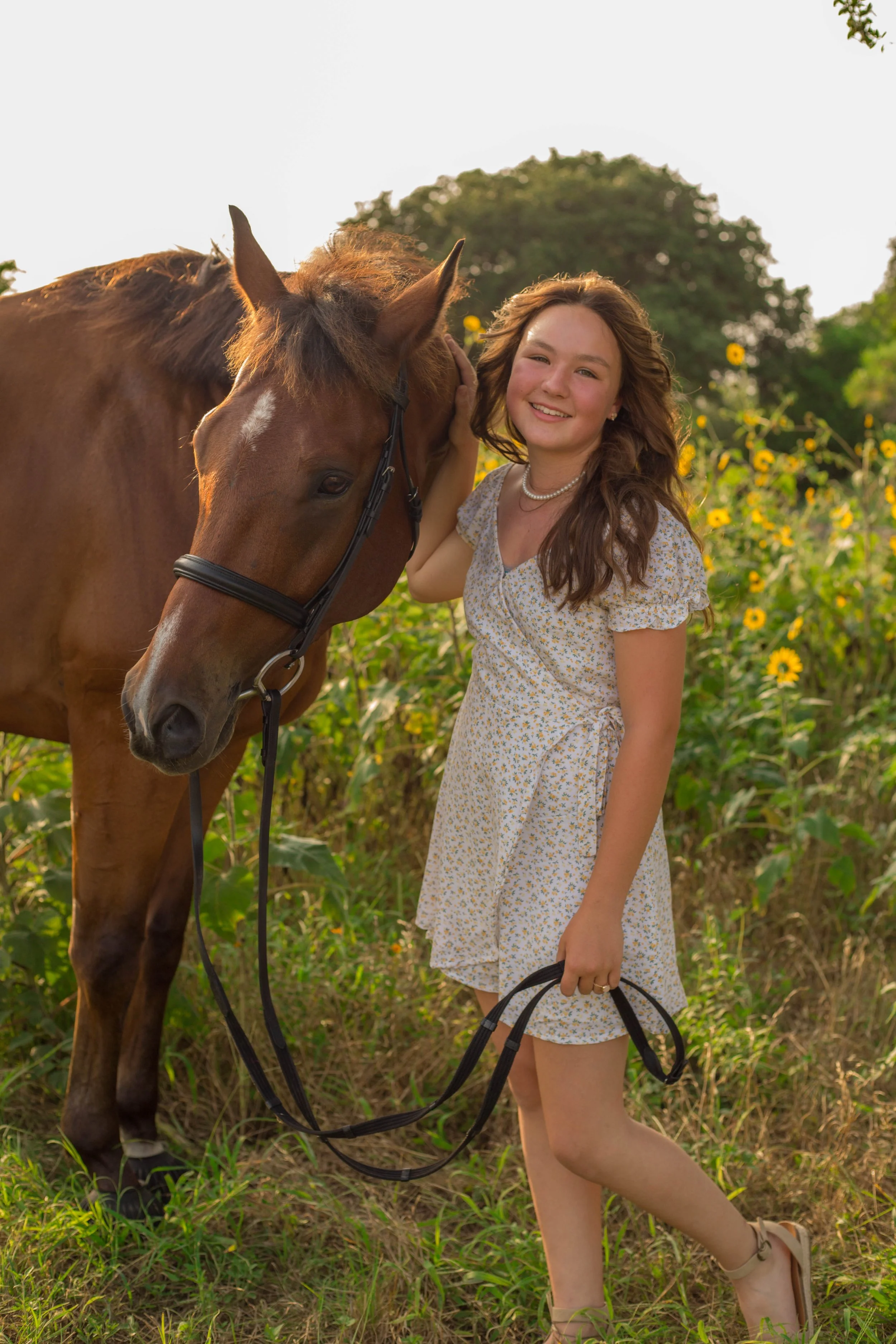 A young woman with long, wavy brown hair wearing a light-colored floral dress with short puffy sleeves, standing outdoors in a field of yellow flowers, smiling and petting a brown horse with a white star on its forehead.