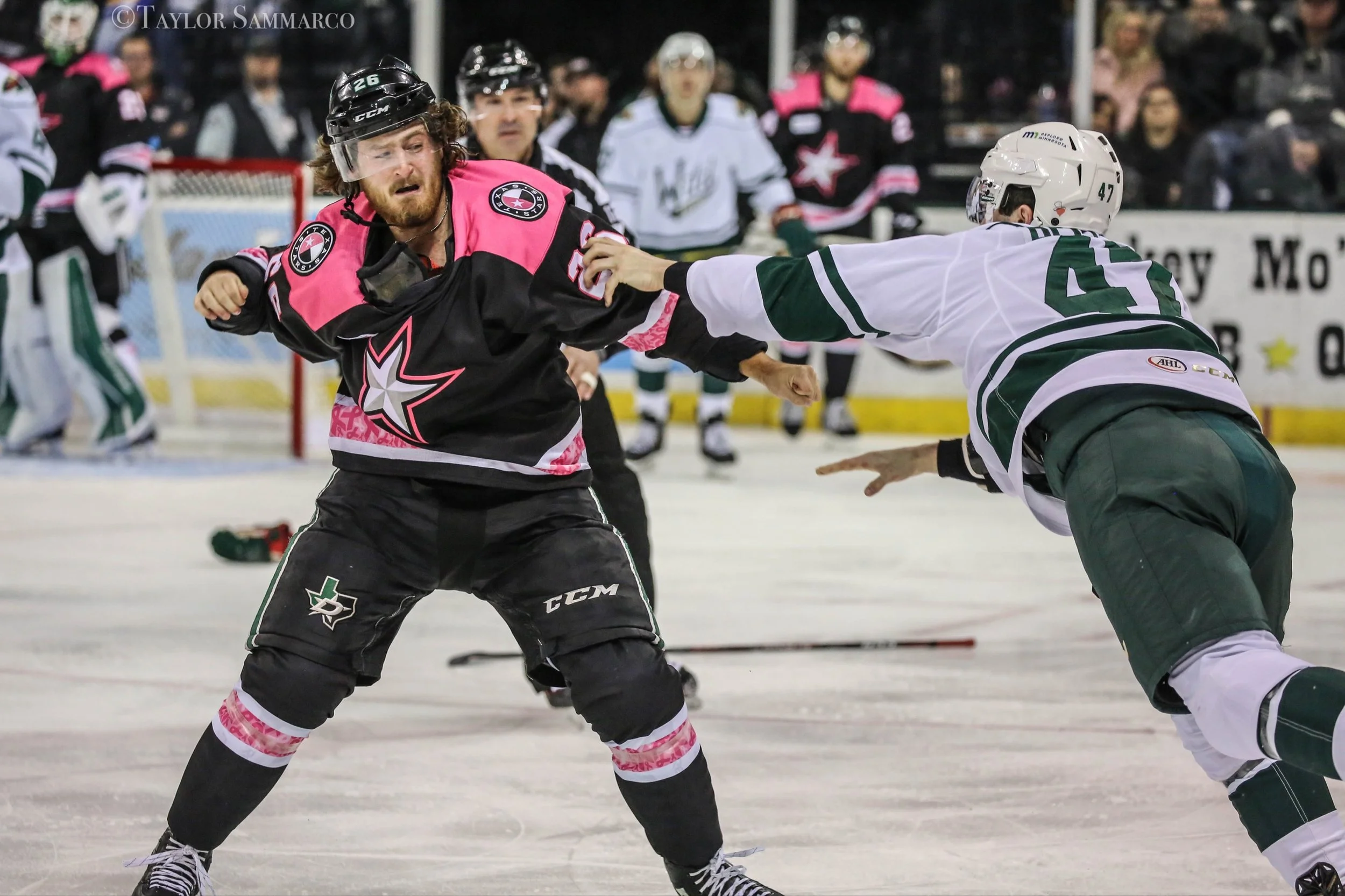 Ice hockey players in action on the ice rink during a game, with one wearing a pink and black jersey and another in a white and green jersey. Spectators in the background.