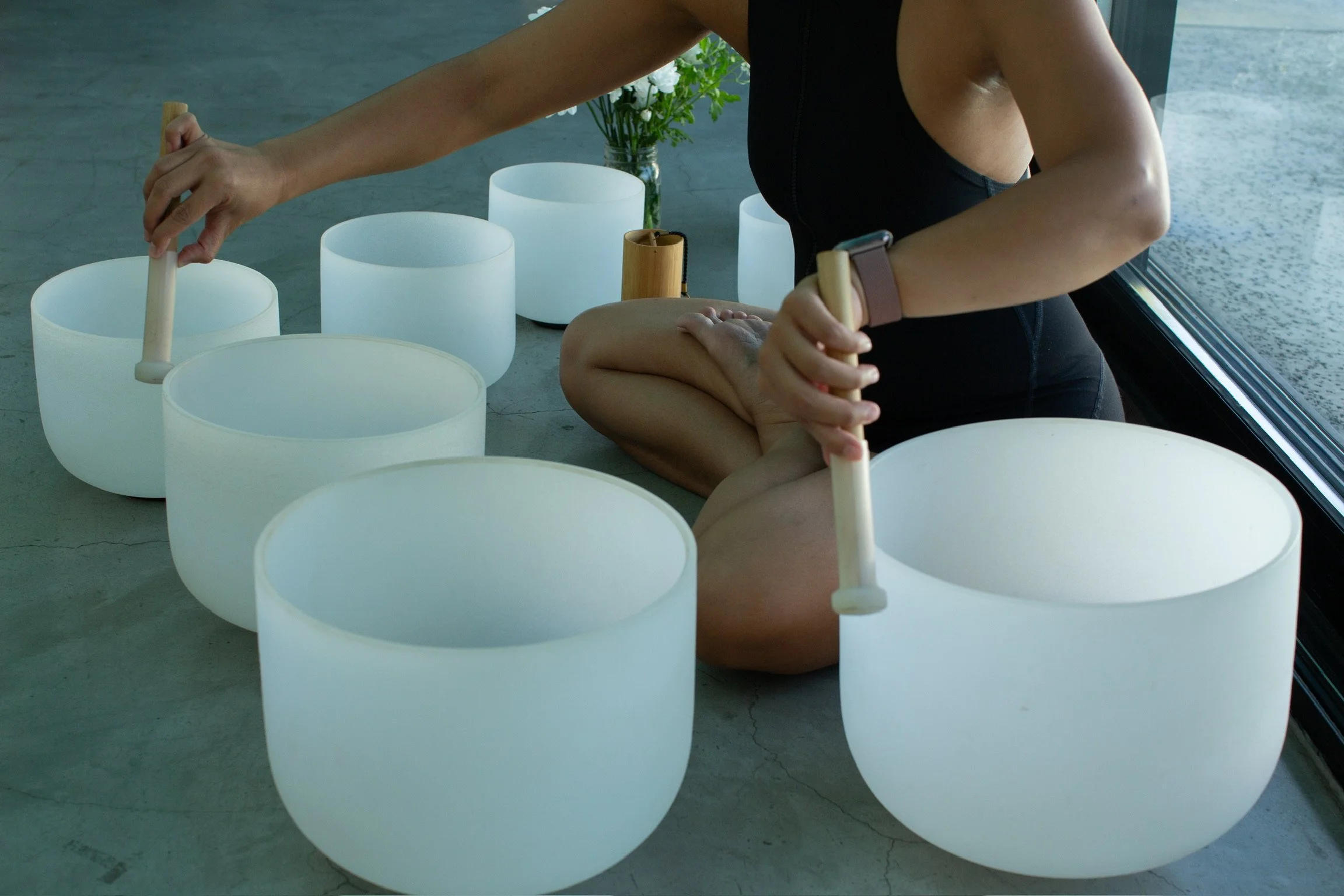 Person sitting cross-legged on the floor playing a wooden percussion instrument, surrounded by white singing bowls and a vase with white flowers.