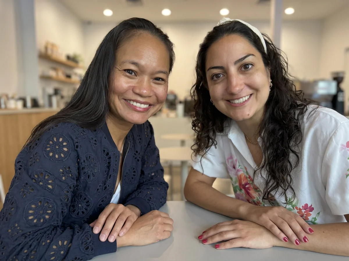 Two women sitting at a table in a bright, modern cafe, smiling at the camera.