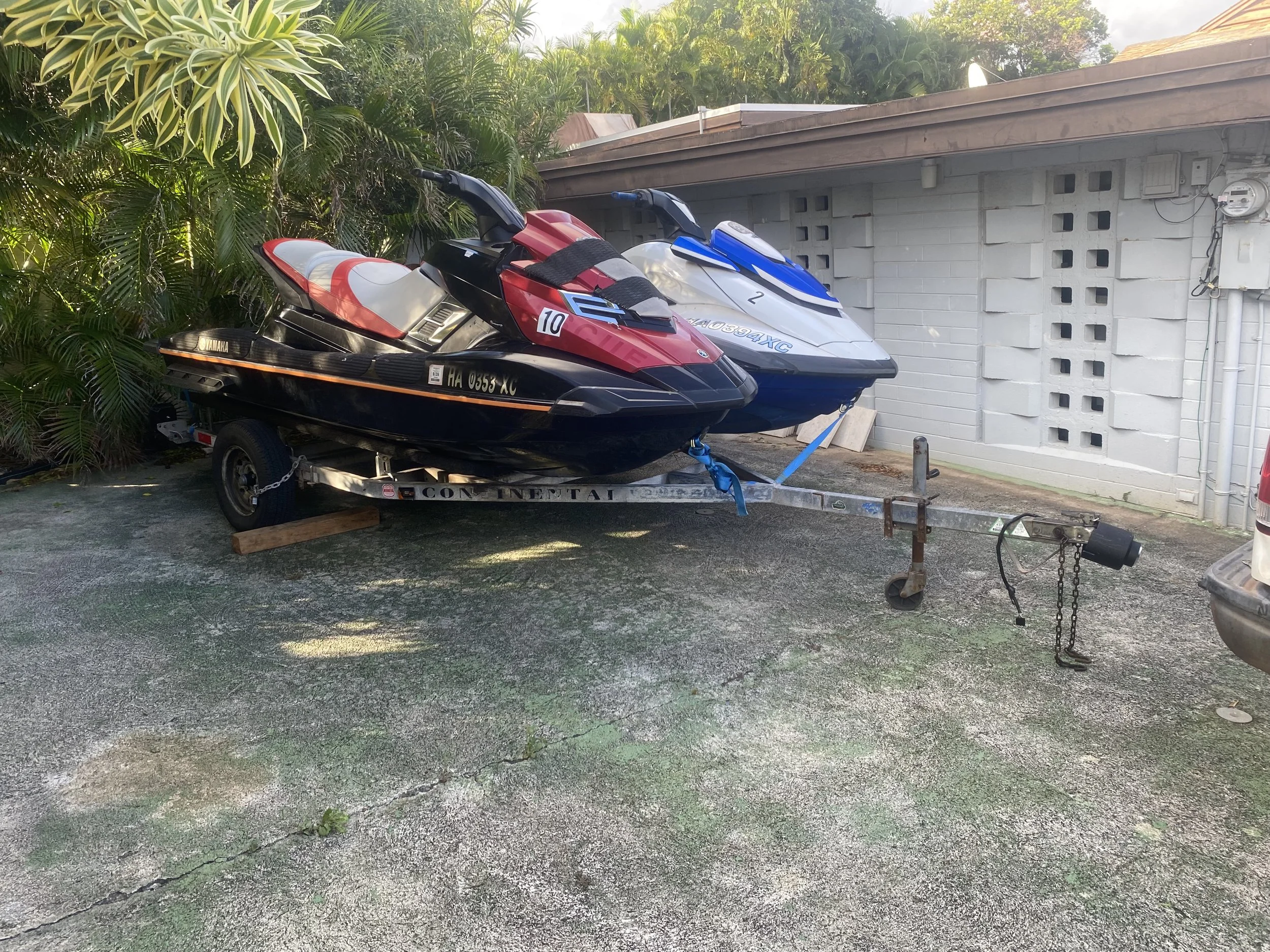 Two jetskis, one black with red and white accents and the number 10, and one blue and white with the number 2, are on a trailer parked on a gravel surface next to a gray wall, with lush green plants behind them. Rental Jet Ski Business in Honolulu 
