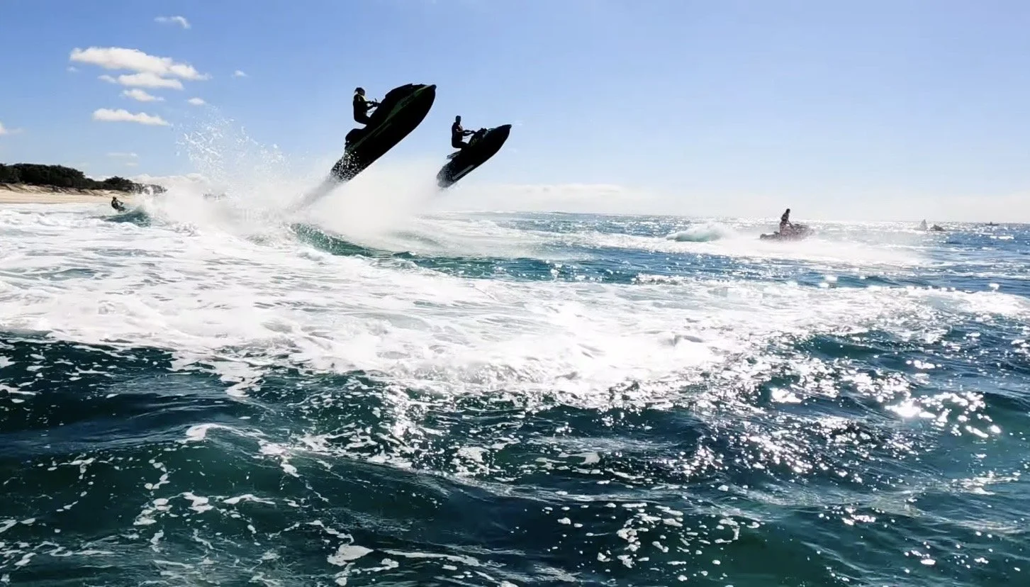 Two people in jet skis jumping out of the water with splashes, while other jet skis are visible in the background, on a sunny day with clear blue sky.