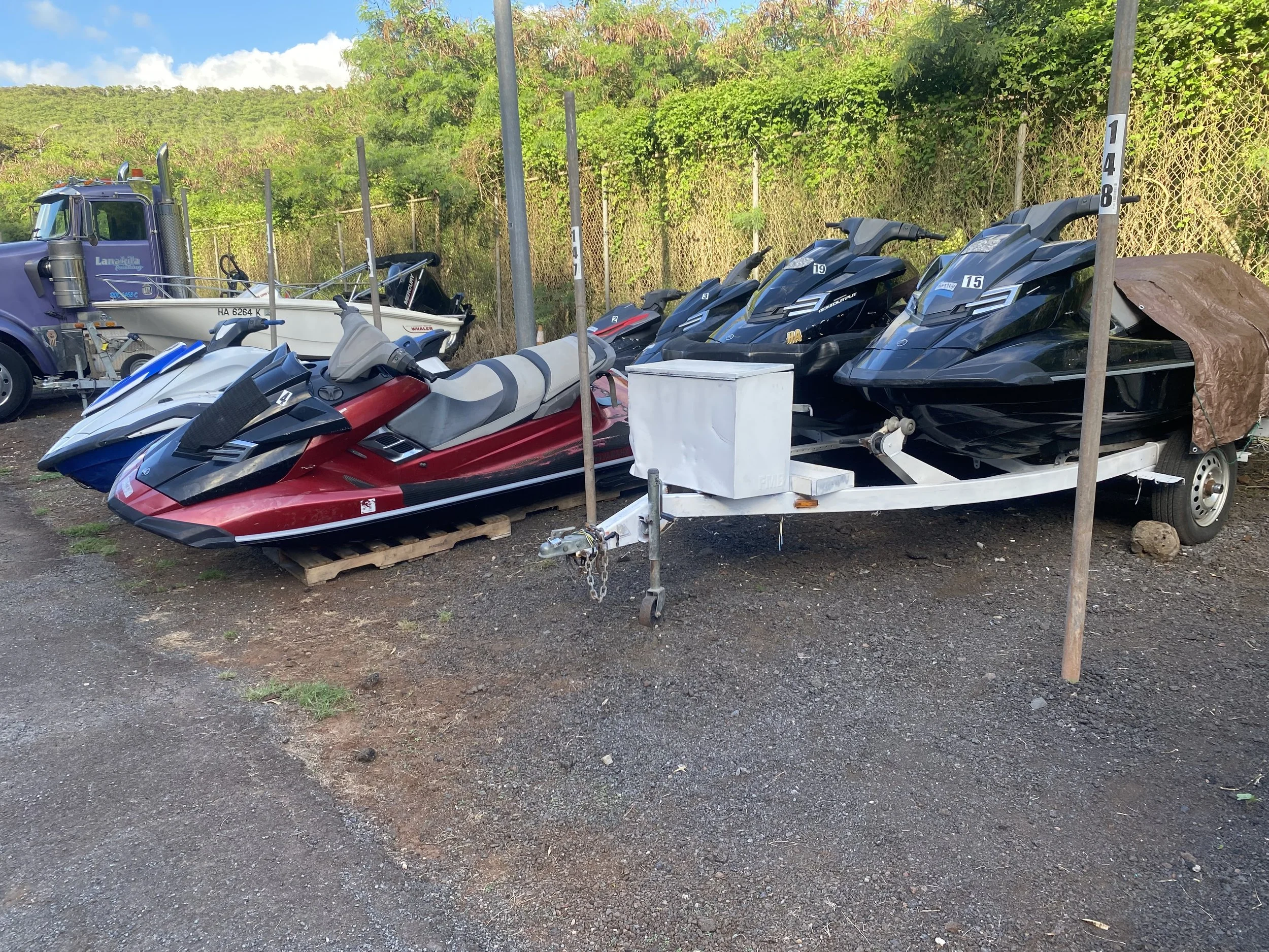 A trailer carrying multiple personal watercraft, including jet skis and a small boat, parked outdoors on a dirt surface with a chain link fence and green trees in the background in Honolulu Hawaii