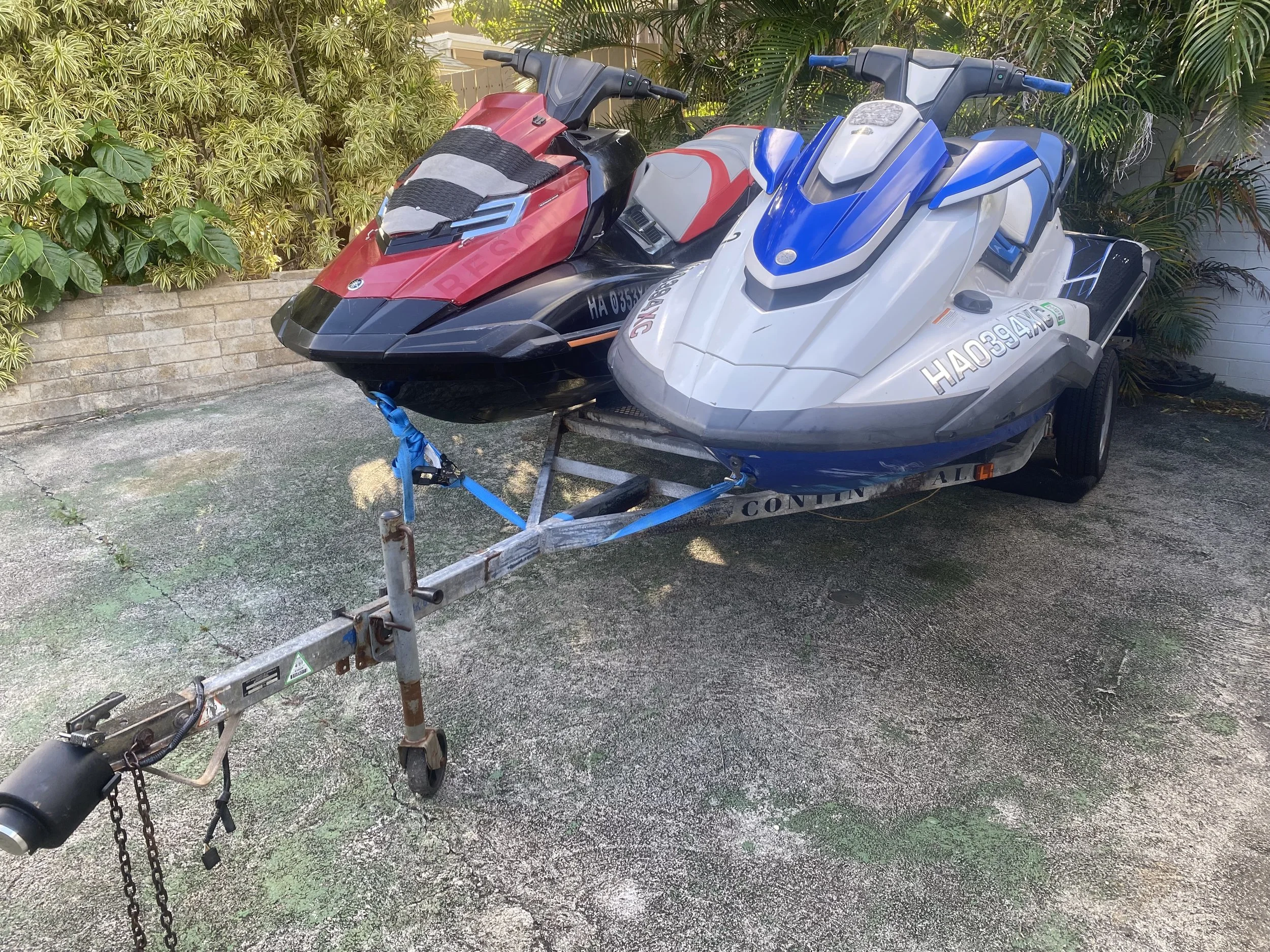 Two jet skis, one red and black and the other white and blue, are on a trailer parked outdoors near plants and a brick wall In Honolulu Hawaii. They are Rentals.