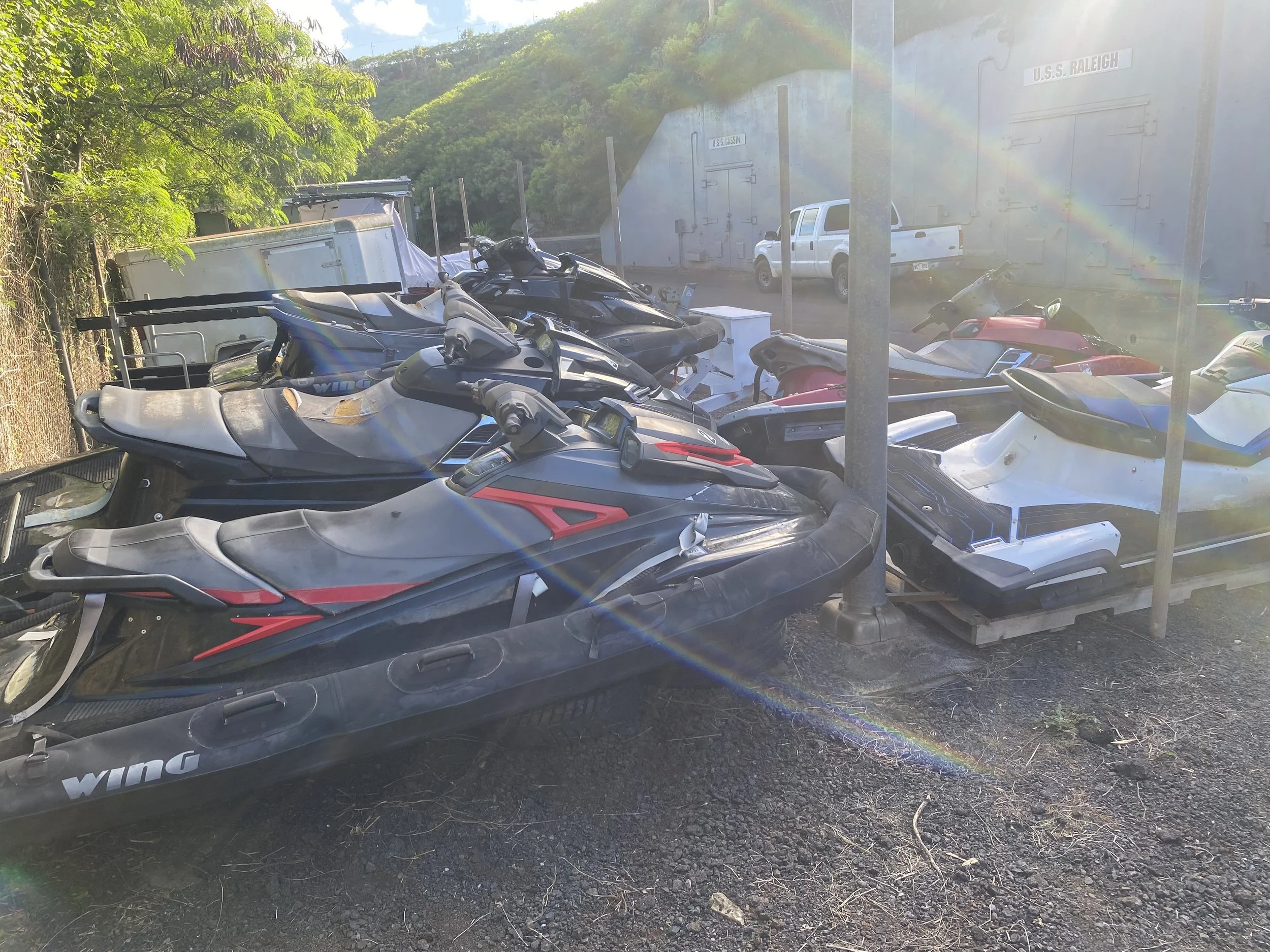 Multiple jet skis loaded on a trailer with trees and a building labeled 'U.S.S. Raleigh' in the background, sunlight creating rainbow flare effects.