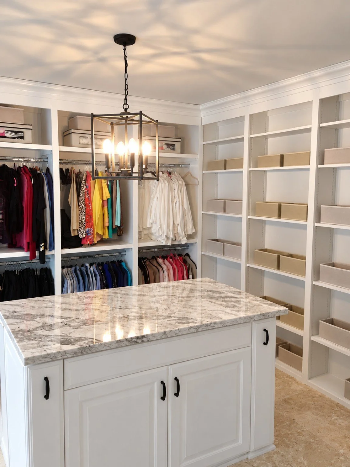 A walk-in closet with white shelves and a marble-topped island in the foreground, containing clothing and storage bins, with a chandelier hanging from the ceiling.