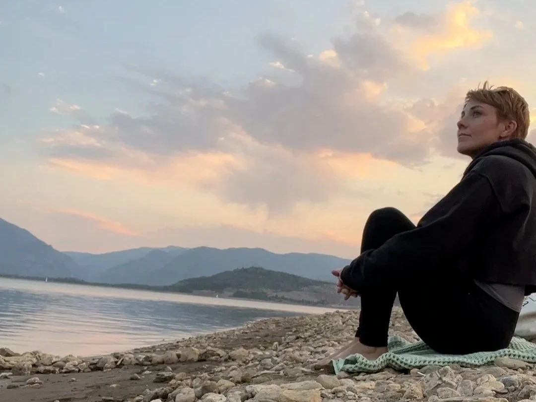 Katie Grace sitting on a rocky beach, looking at the water and distant mountains during sunset.