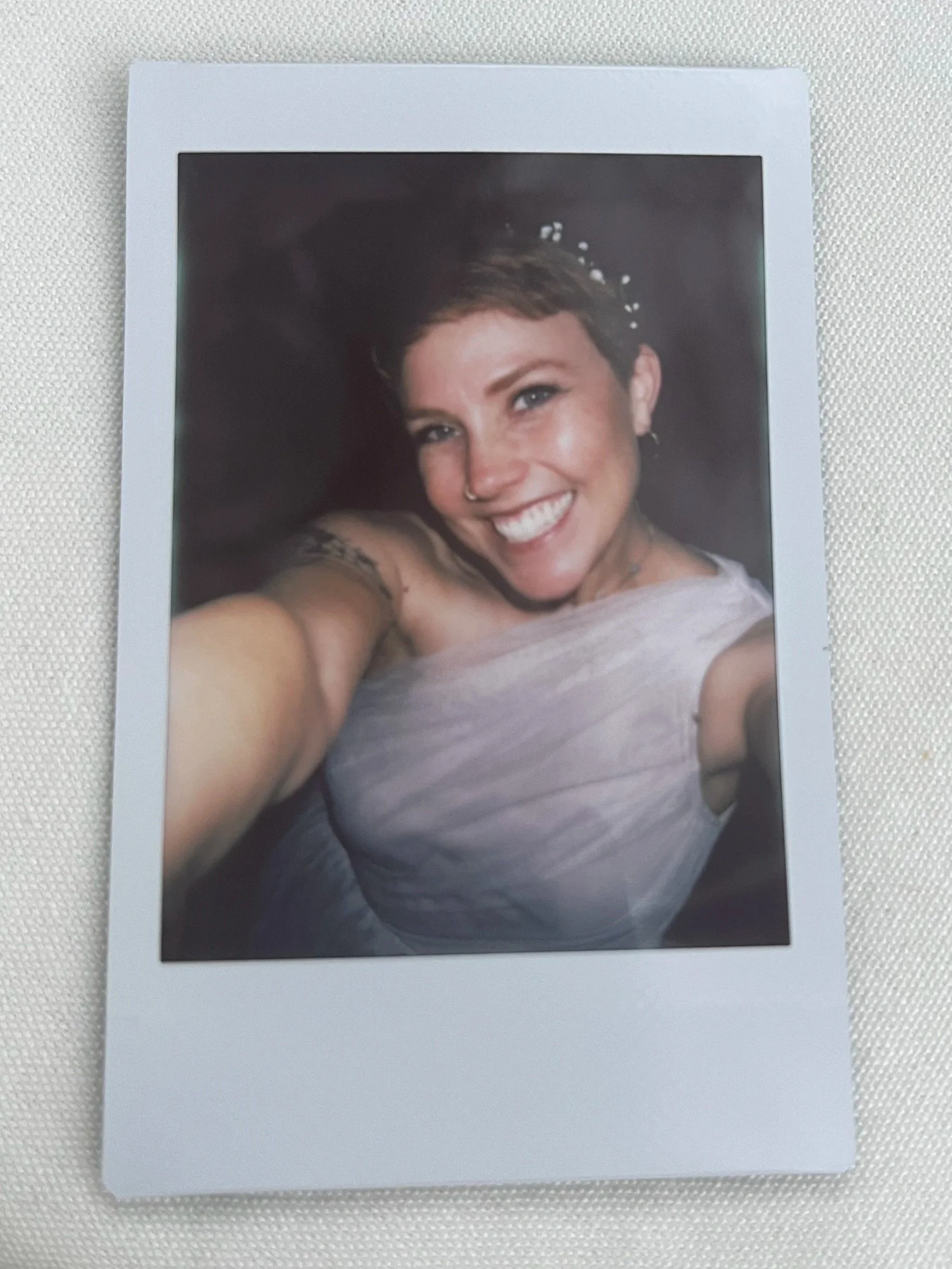 A printed photograph of Katie Grace smiling with short brown hair, wearing a rose-colored top, taking a selfie. The photo is against a dark background, and she is smiling widely.