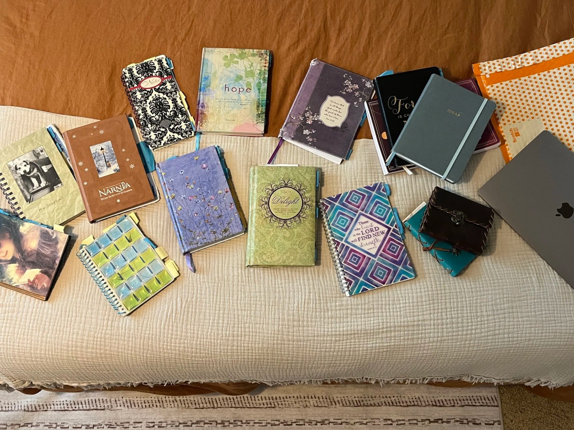 A variety of notebooks, journals, and planners are spread across a beige table with a textured tablecloth, some with colorful covers and decorative patterns.