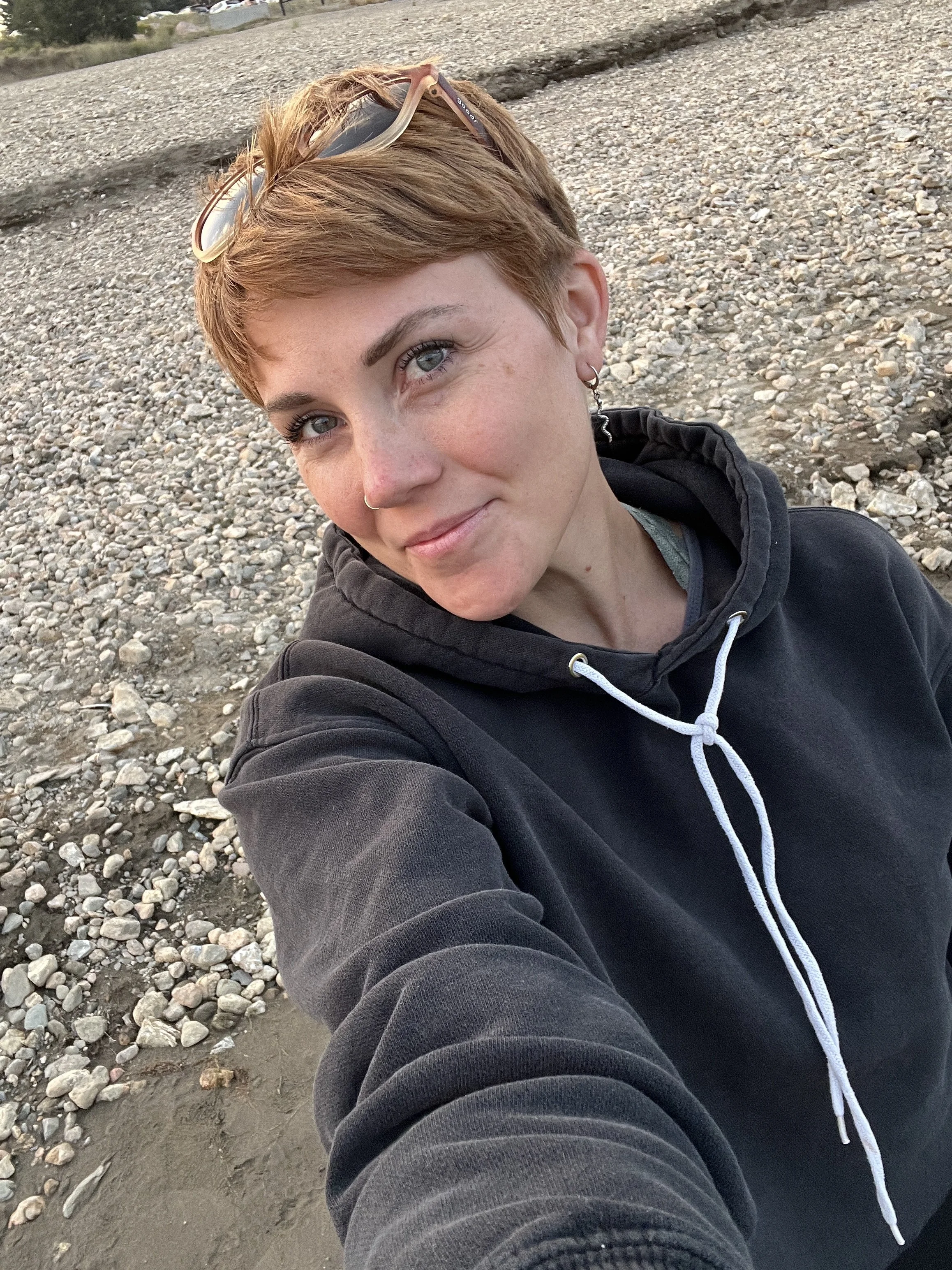 A woman (Katie Grace) with short, light brown hair and blue eyes takes a selfie on a rocky and muddy beach. She is wearing a black hoodie with white drawstrings, earrings, and glasses resting on her head.