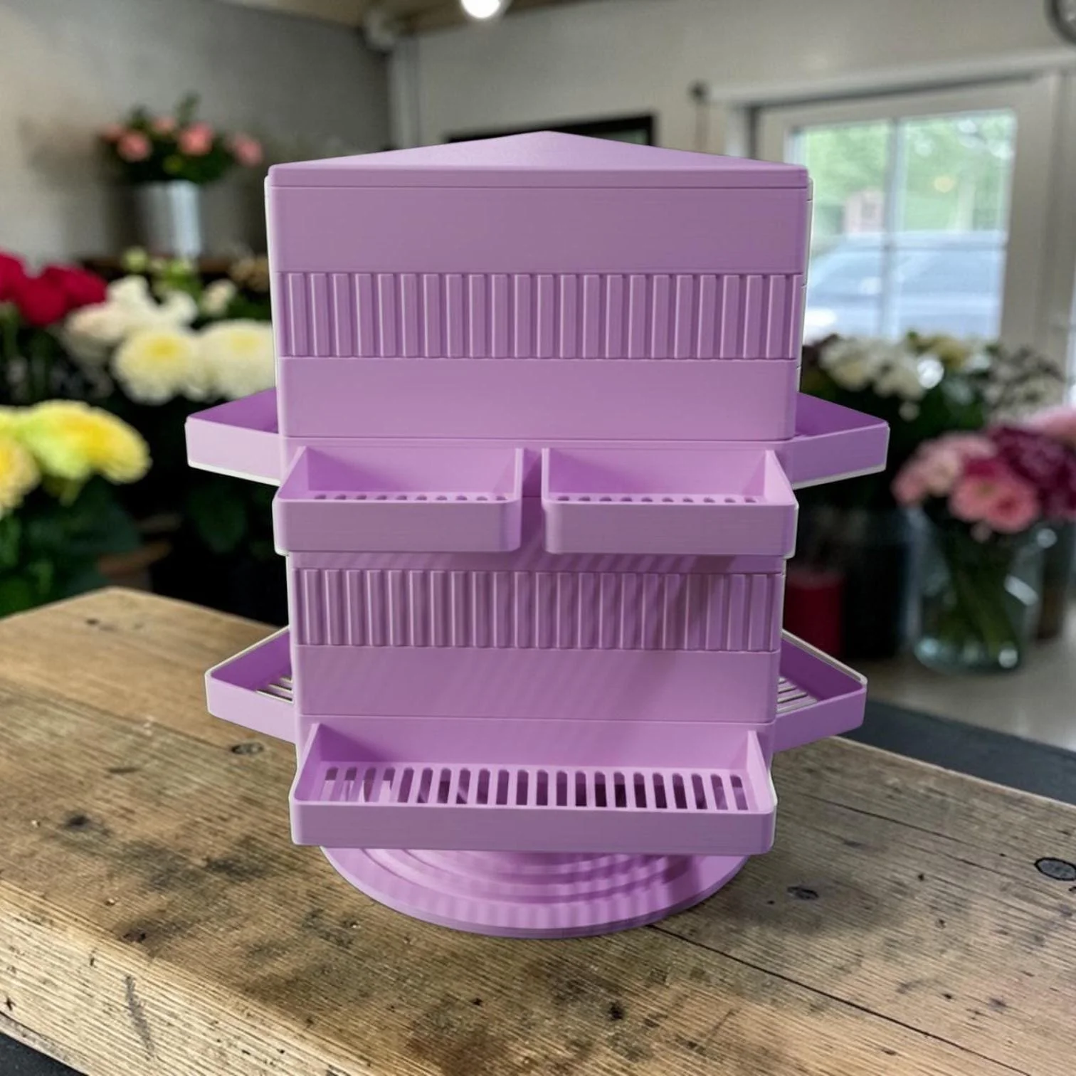 A pink plastic multi-tiered storage organizer with multiple compartments, placed on a wooden surface in a room with flowers and a window in the background.