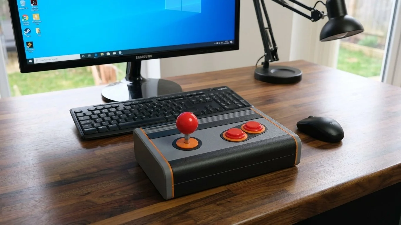 Retro arcade-style gaming setup with a monitor, keyboard, mouse, desk lamp, and a vintage style game controller with a joystick and two action buttons on a wooden desk.