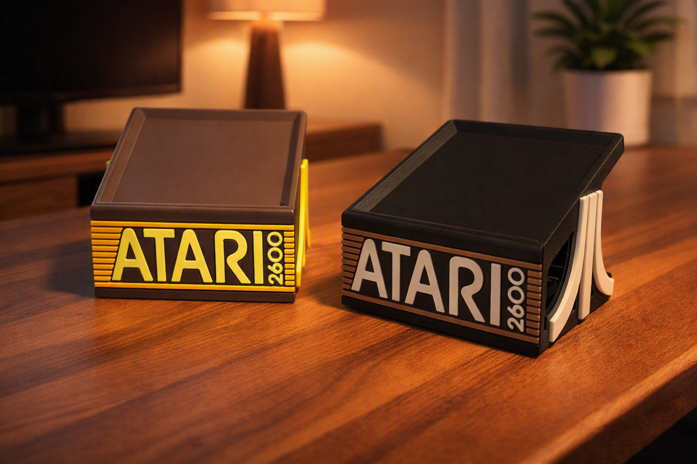 Two vintage Atari 2600 gaming consoles on a wooden surface, one with yellow accents and the other with white accents, in a warmly lit room.