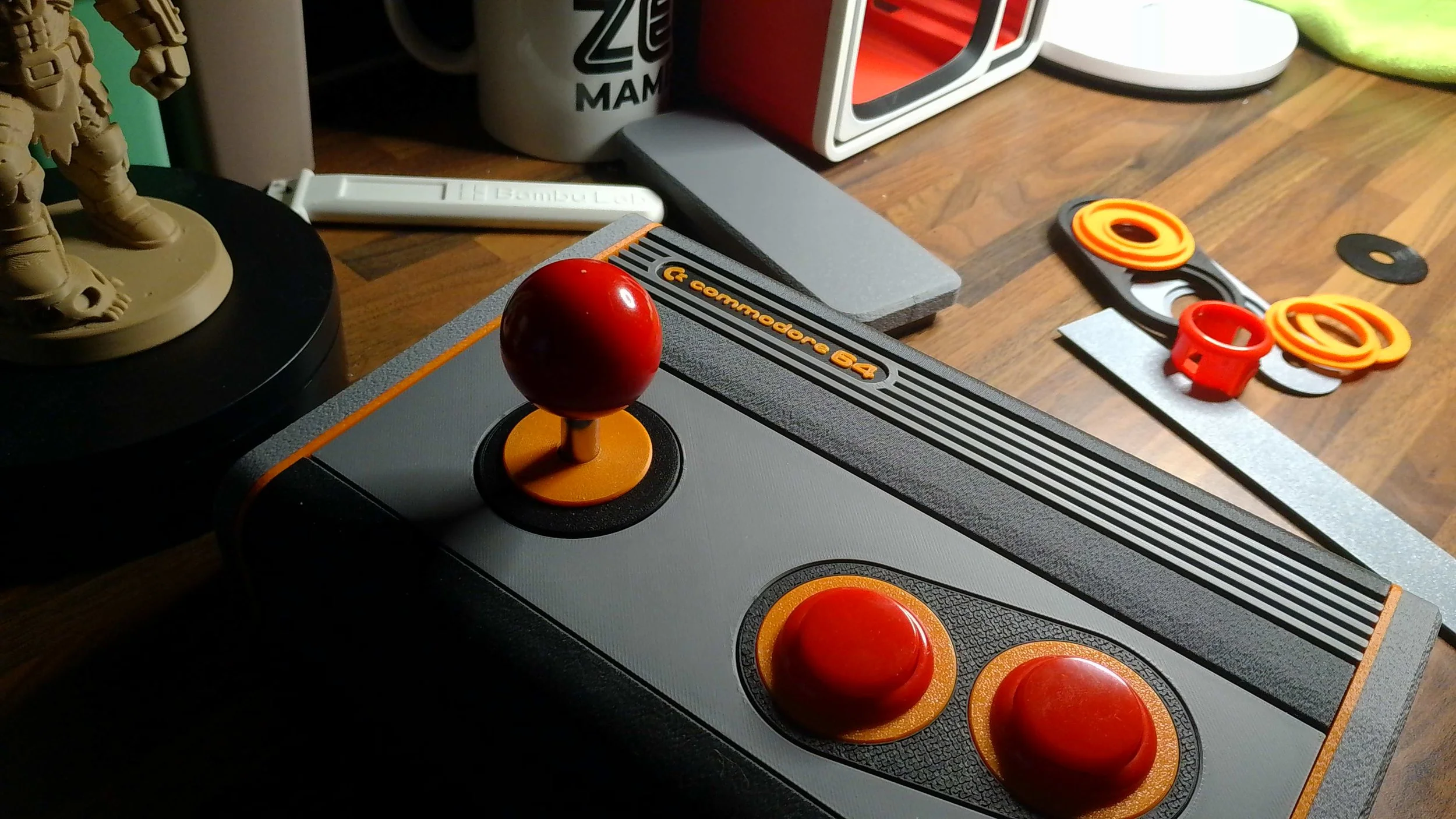 Close-up of a vintage Atari joystick with red buttons, a red ball handle, and a gray base, on a wooden surface with other vintage gaming accessories and décor items in the background.