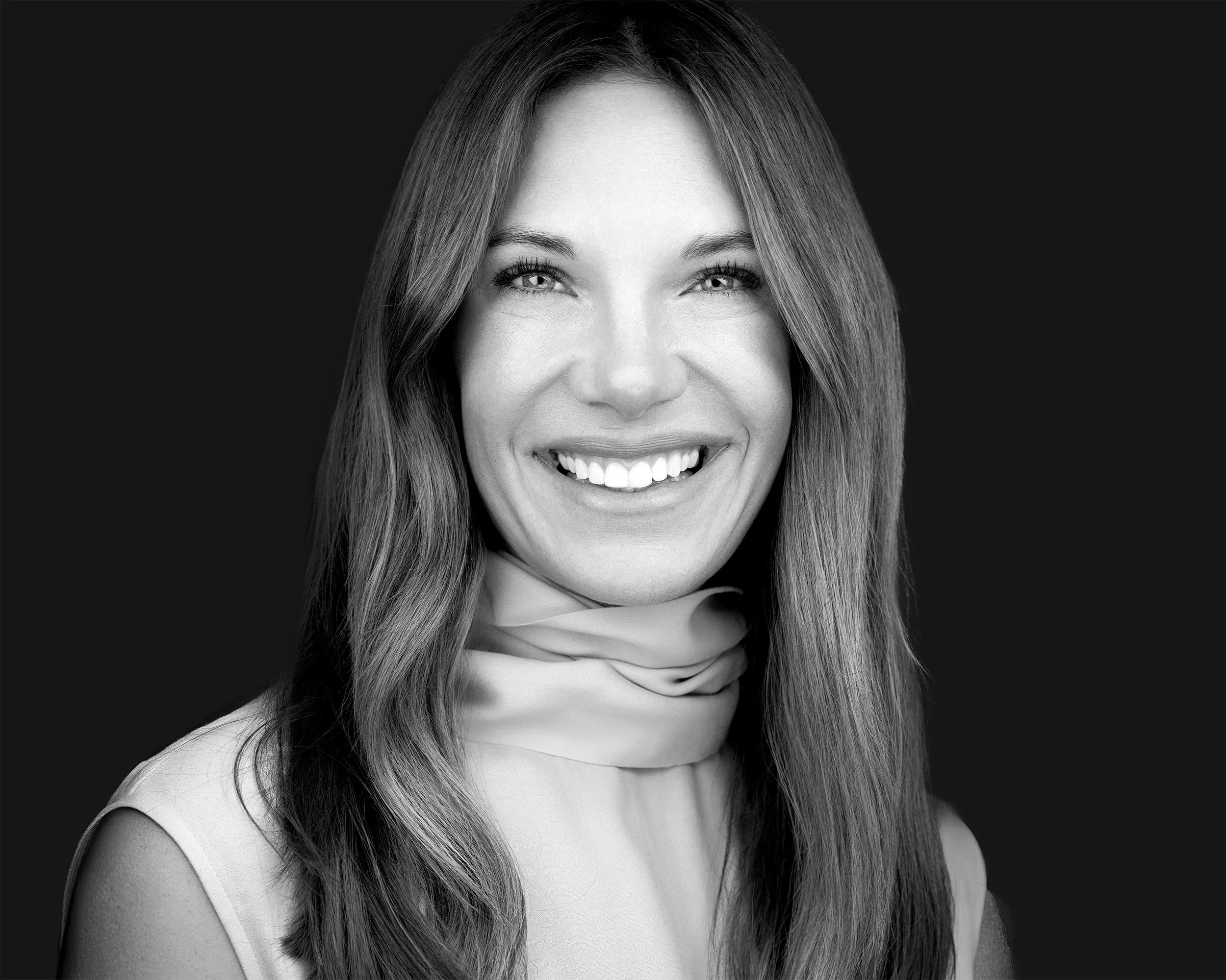 A smiling woman with long hair and a sleeveless top, facing the camera, in black and white, with a dark background.