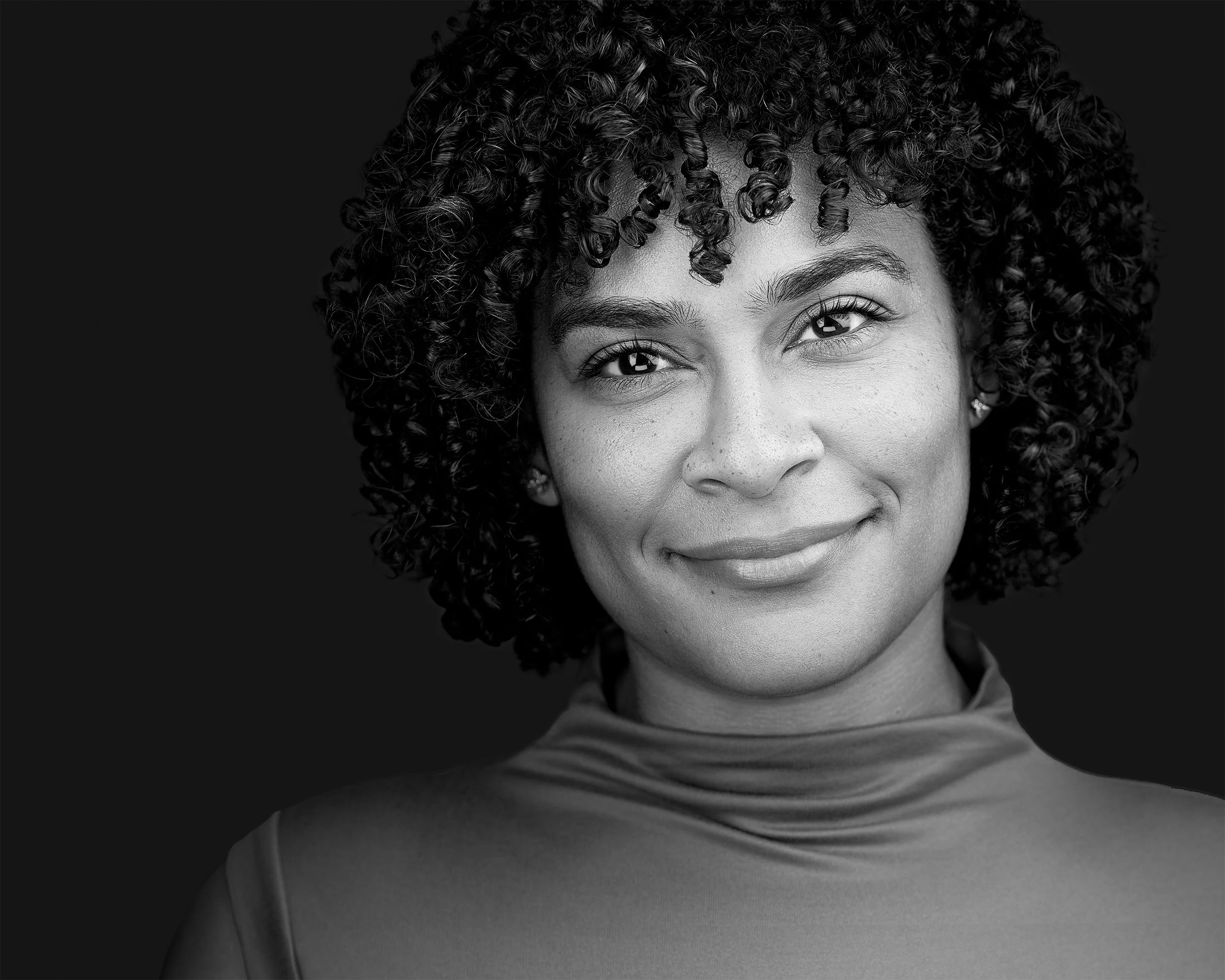 Close-up black and white photo of a smiling woman with curly hair and light skin, wearing a high-neck shirt.