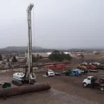 A tall construction crane is lifting a large object in a construction site with vehicles and buildings in the background, under a cloudy sky.