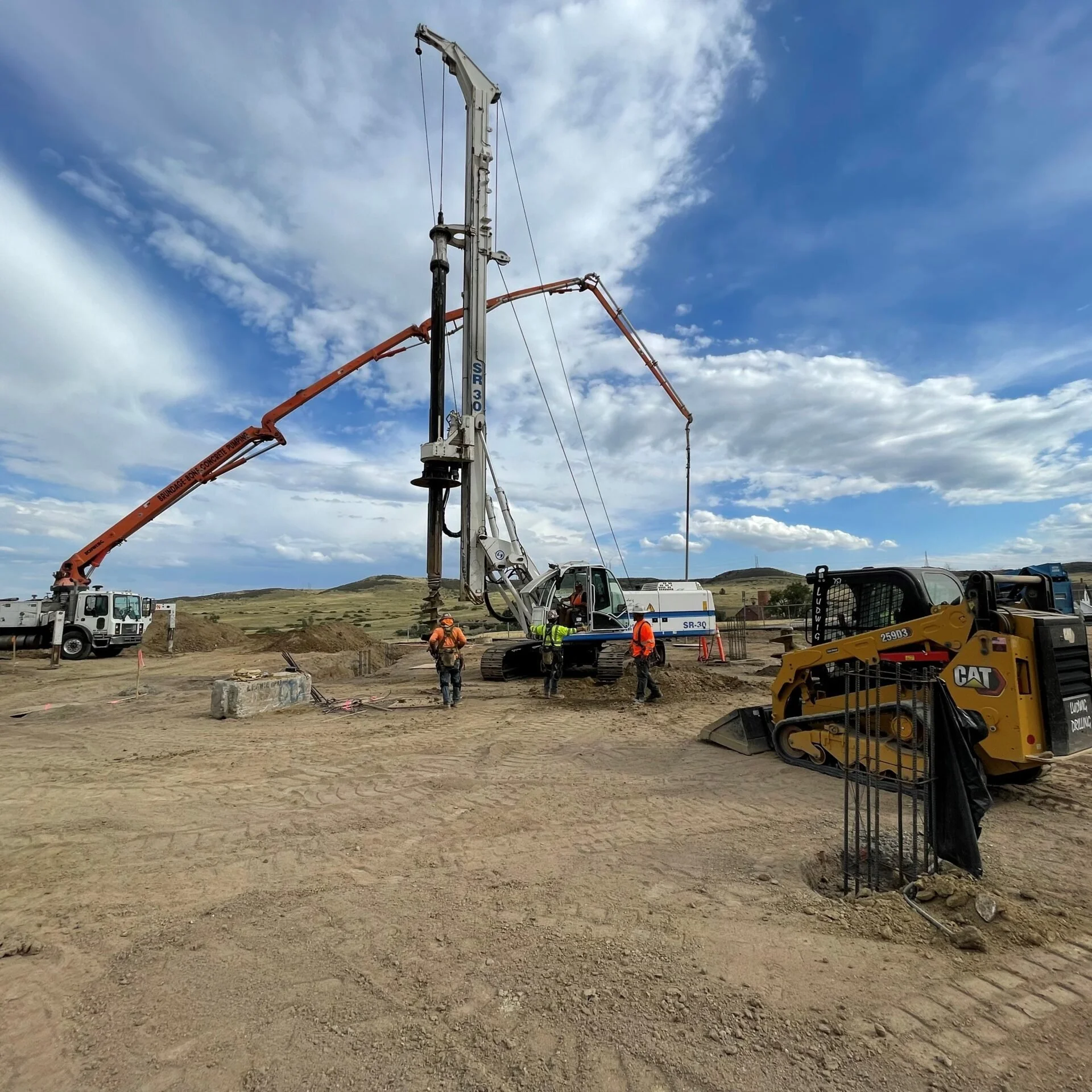 Construction site with heavy machinery, workers in safety vests and helmets, and an overcast sky.