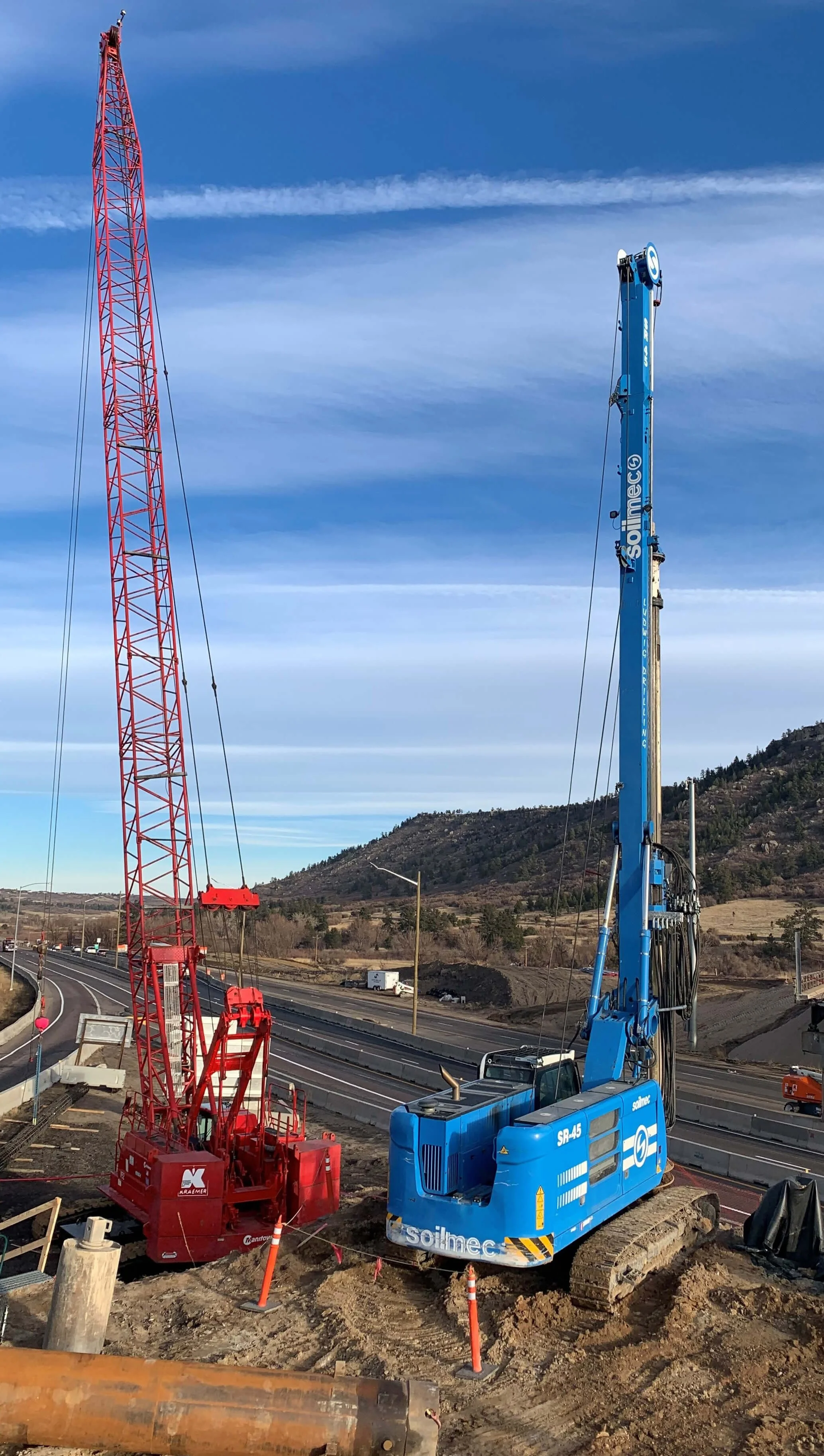 Construction site with red and blue cranes over a highway. Mountains in the background under a blue sky.