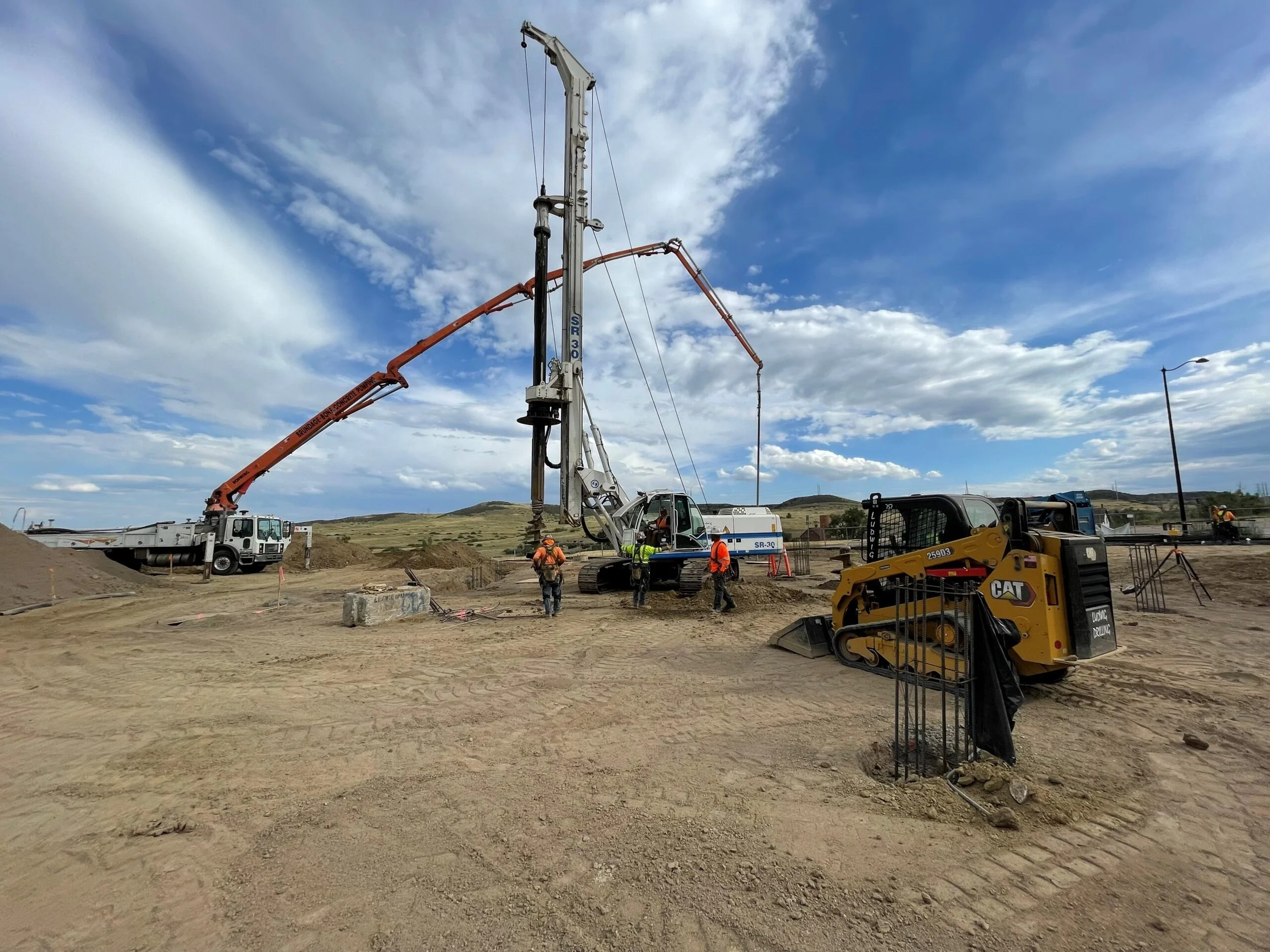Construction site with workers, a concrete pump truck, and heavy machinery on dirt ground under a partly cloudy sky.
