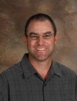 A man with short dark hair, wearing glasses and a gray checked shirt, smiling against a brown background.