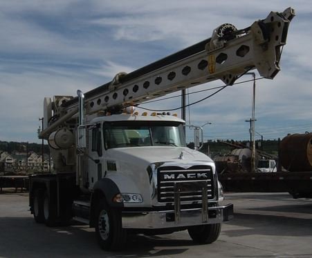 A white Mack truck with a large boom lift attachment on the back, parked in an industrial area.