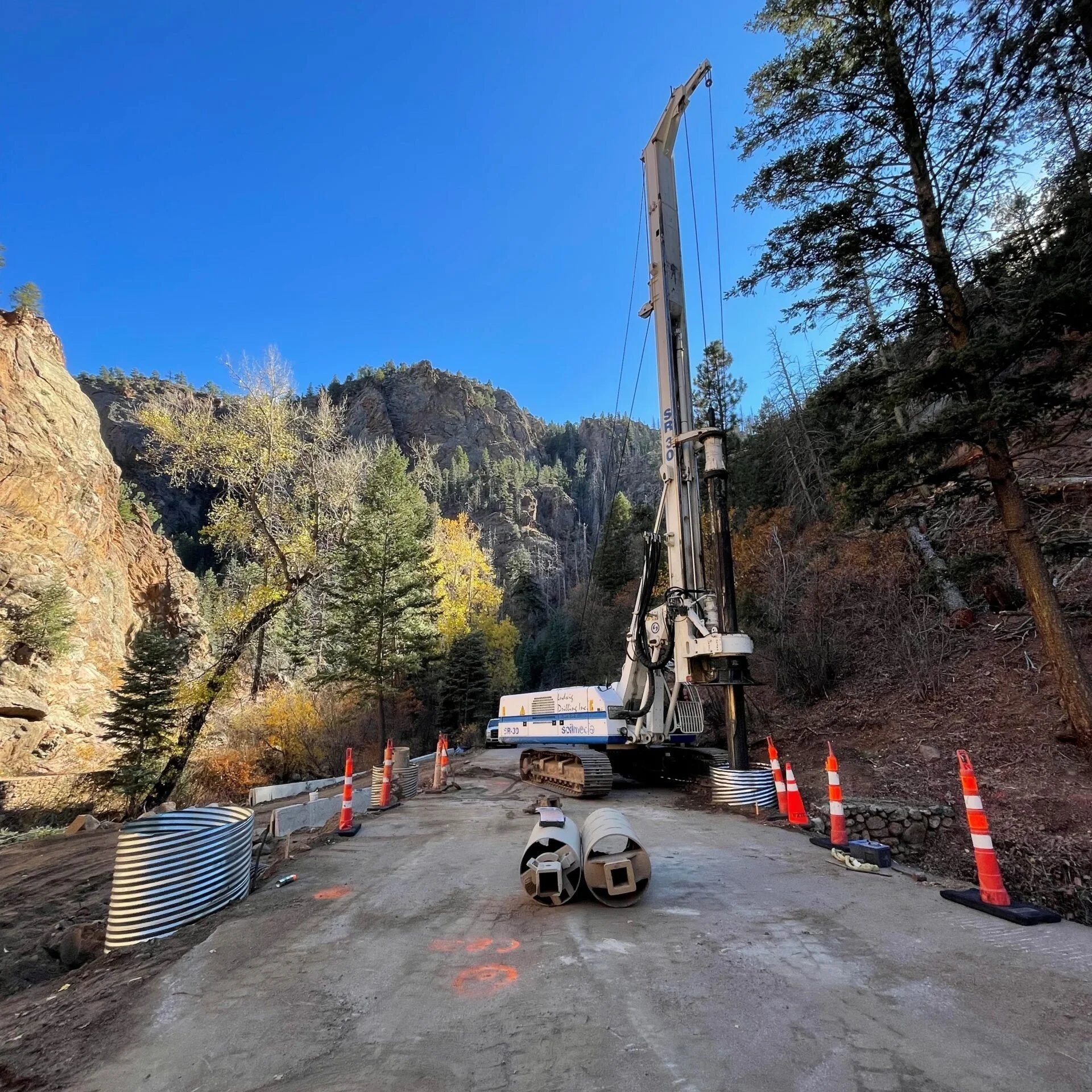 Construction site on a mountain road with orange traffic cones, metal drainage pipes, and a large construction drilling machine against a backdrop of rocky mountains, trees, and a clear blue sky.
