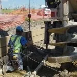 Construction workers in safety gear working at a construction site with orange safety fencing and construction equipment.
