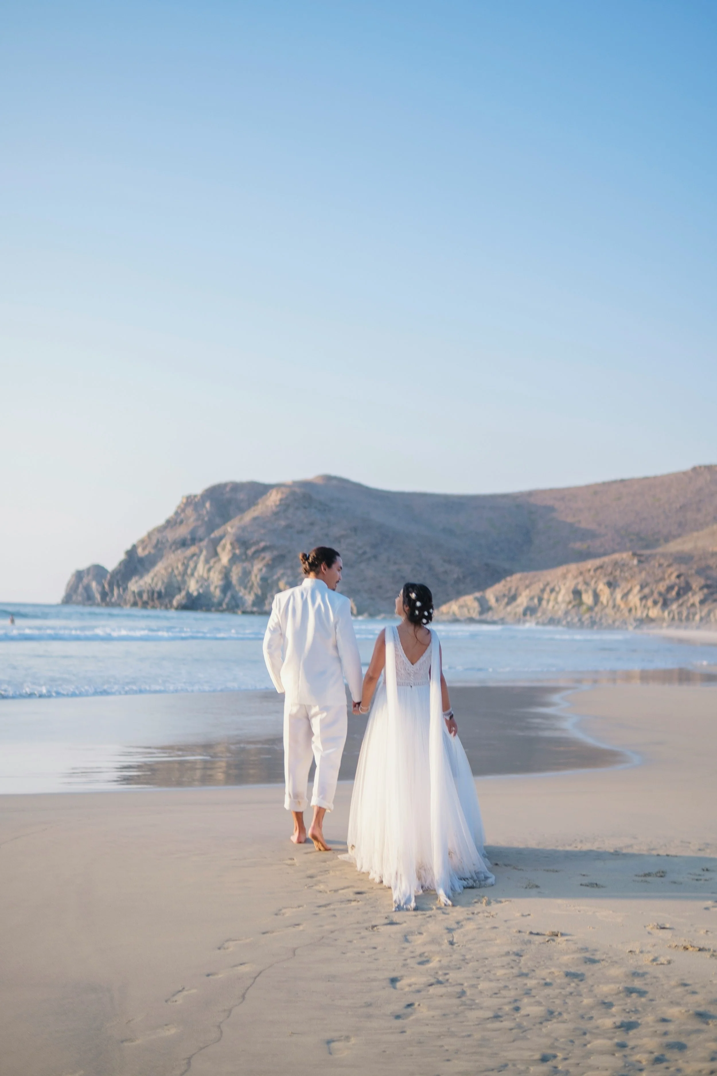 A couple dressed in wedding attire walking hand-in-hand along the beach at sunset with a mountainous island in the background.