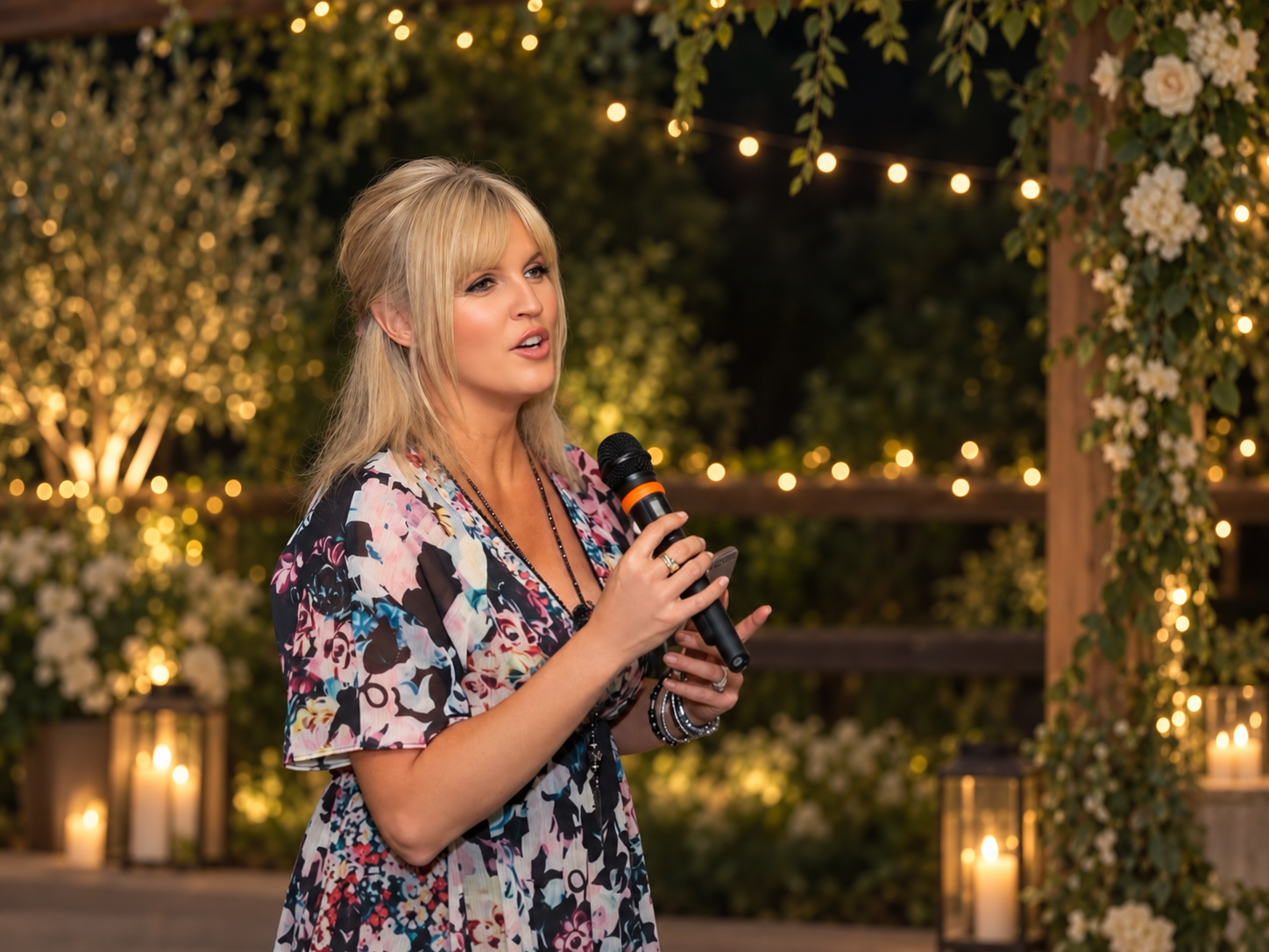 A woman with blonde hair holding a microphone, speaking at an outdoor event decorated with string lights, candles, and flowers.