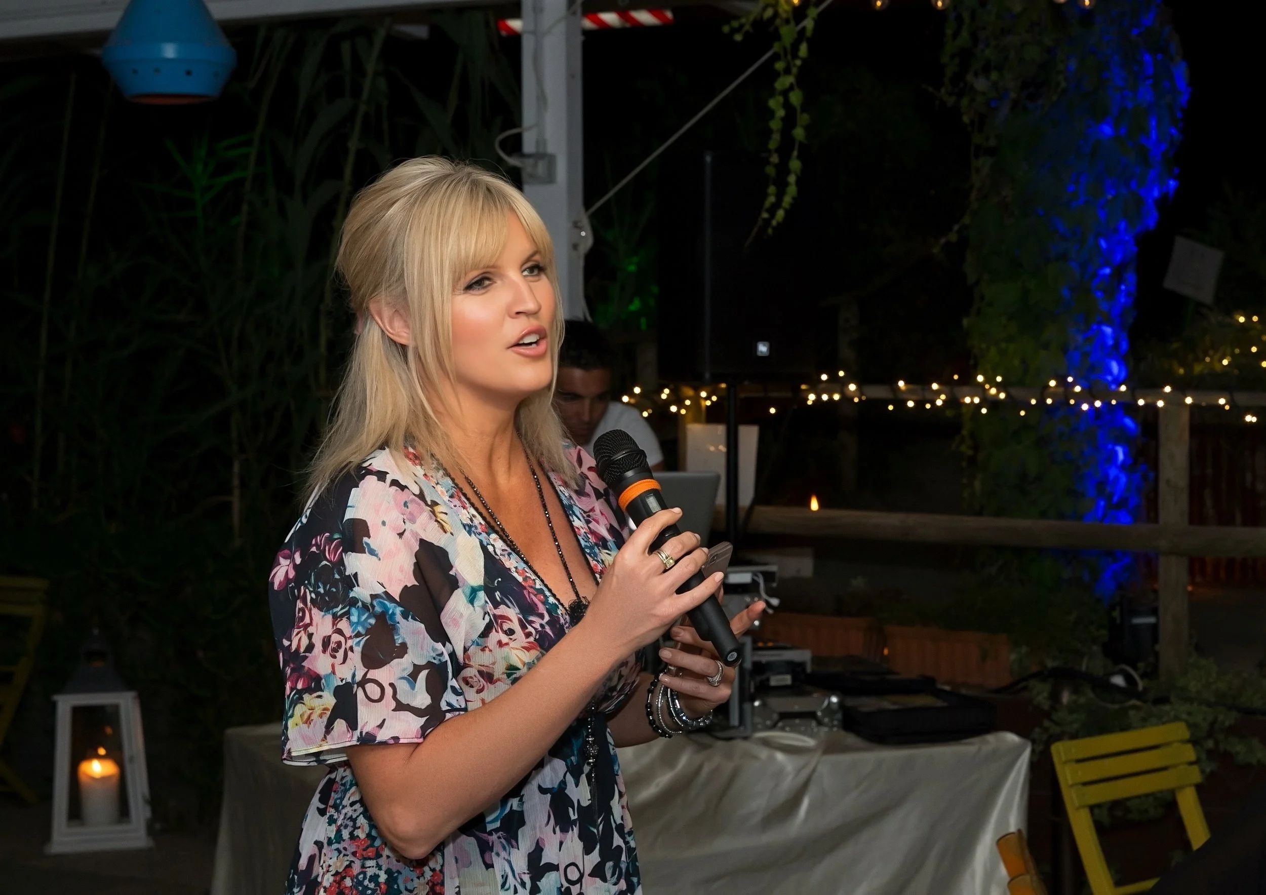 A woman with blonde hair holding a microphone at an outdoor evening event, with string lights, greenery, and decorative lanterns in the background.