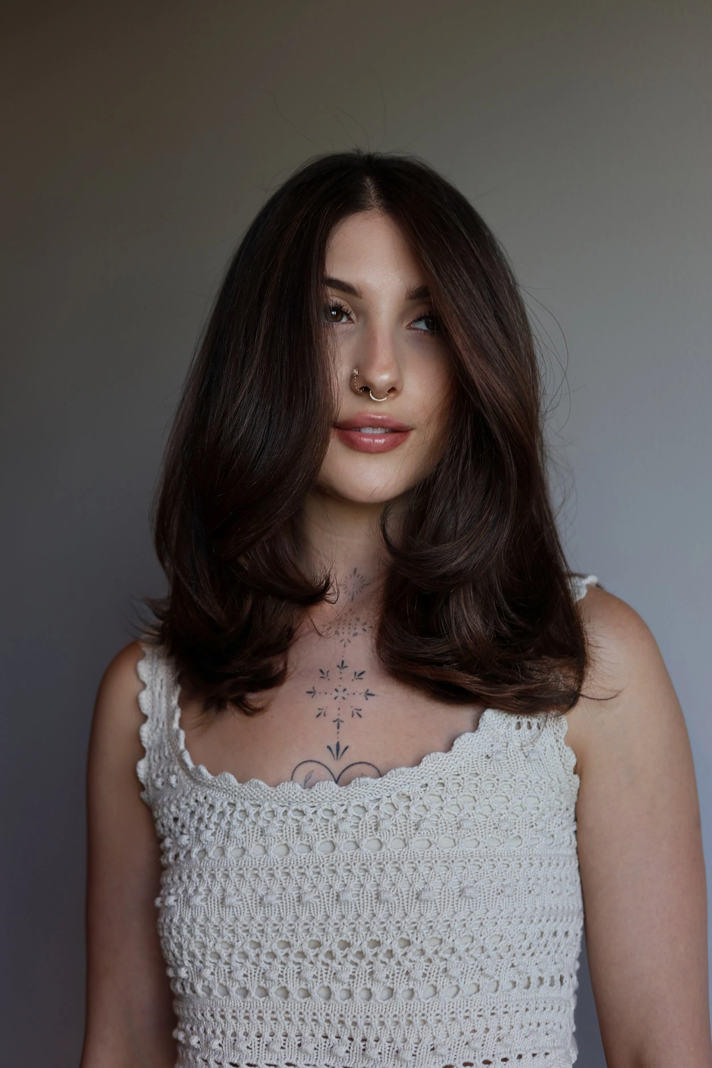 A young woman with long dark hair, light skin, and a septum piercing, wearing a white crocheted top, standing against a plain gray background.