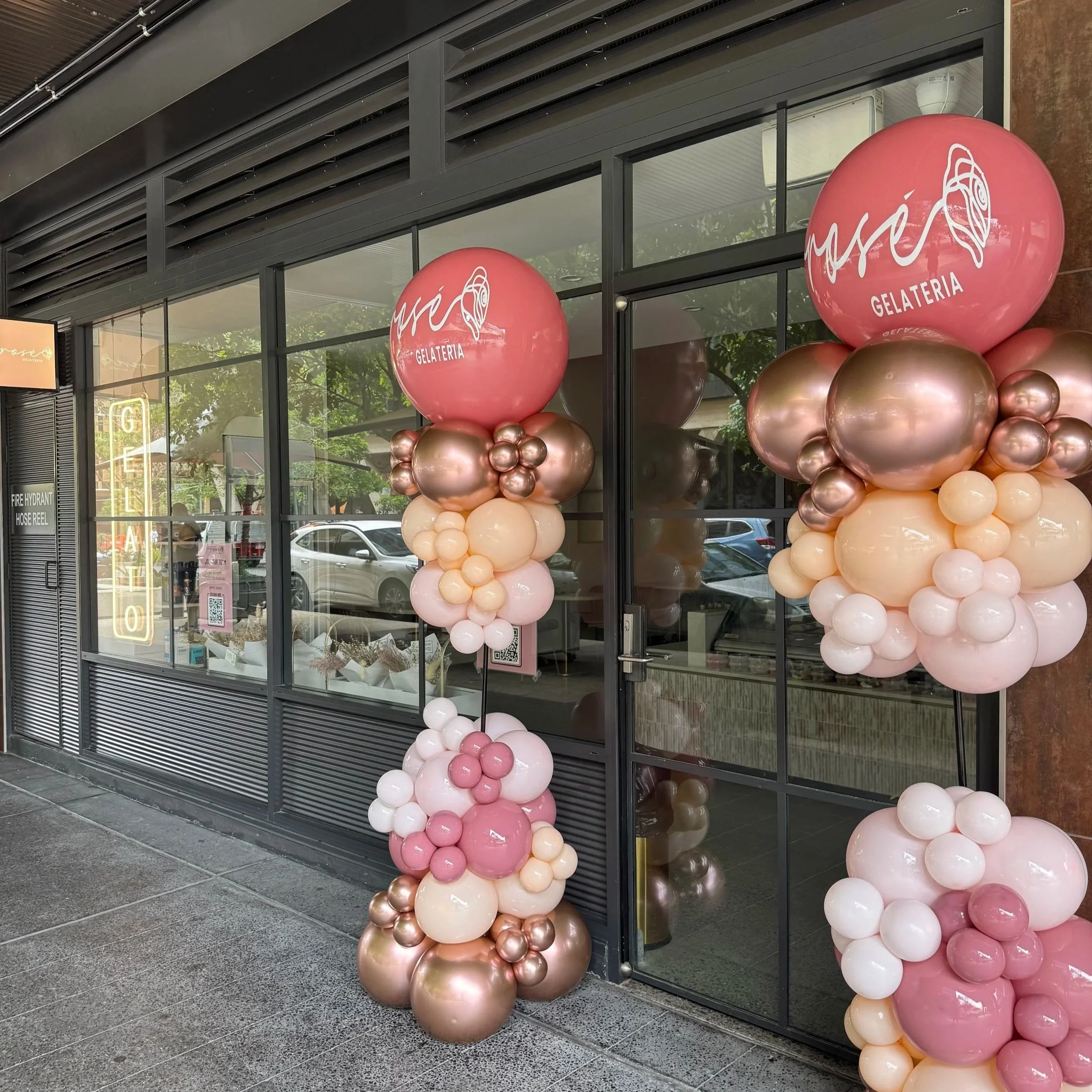 Decorative balloon columns outside a storefront with glass windows, labeled 'MSO GELATERIA'. Large pink and white balloons are arranged in columns on each side of the entrance.