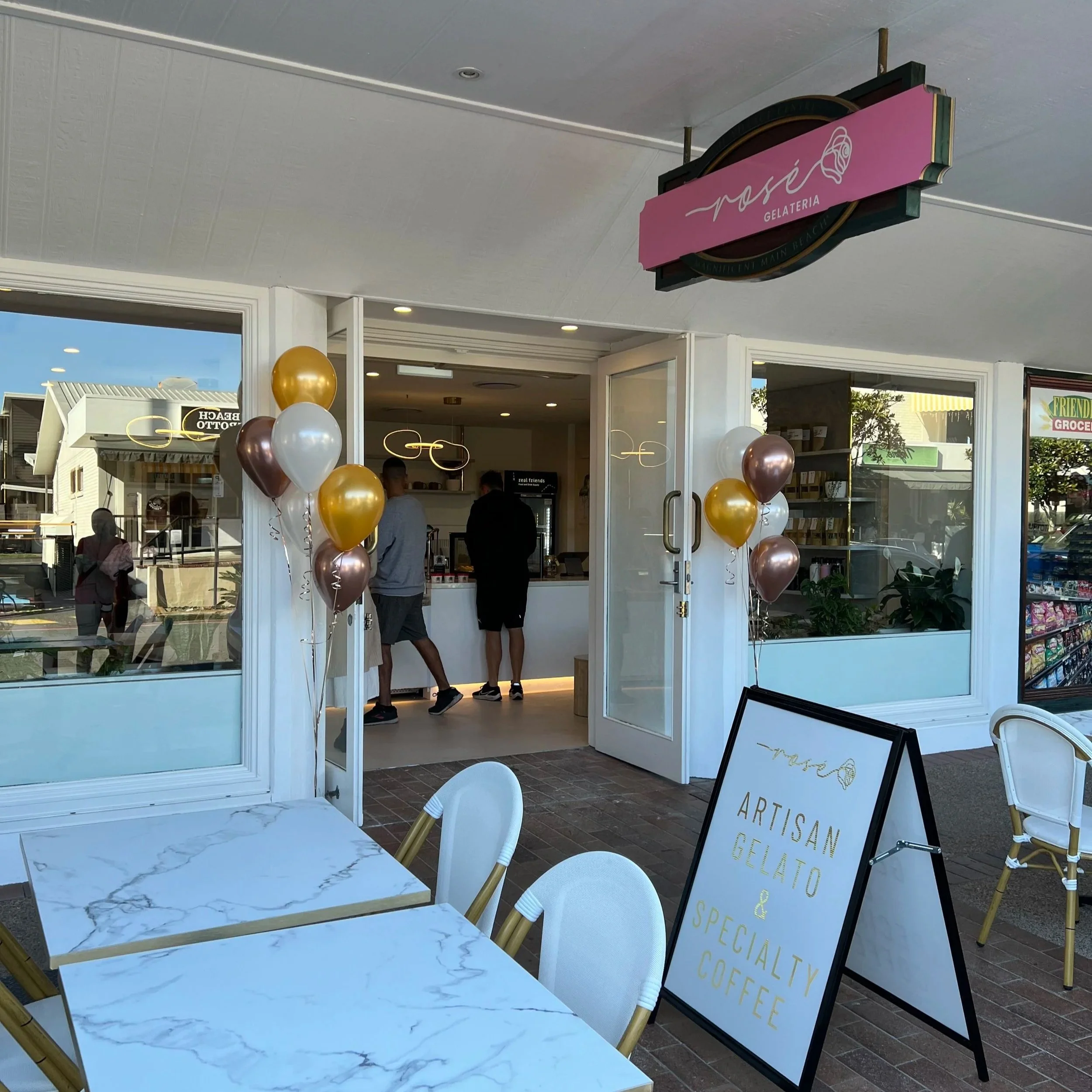 Outside view of a gelato shop with a pink signboard displaying 'nise Gelateria'. Balloons are arranged near the entrance, and inside, a few people are visible. There are chairs and tables for customers, and a black signboard with menu items is also visible.