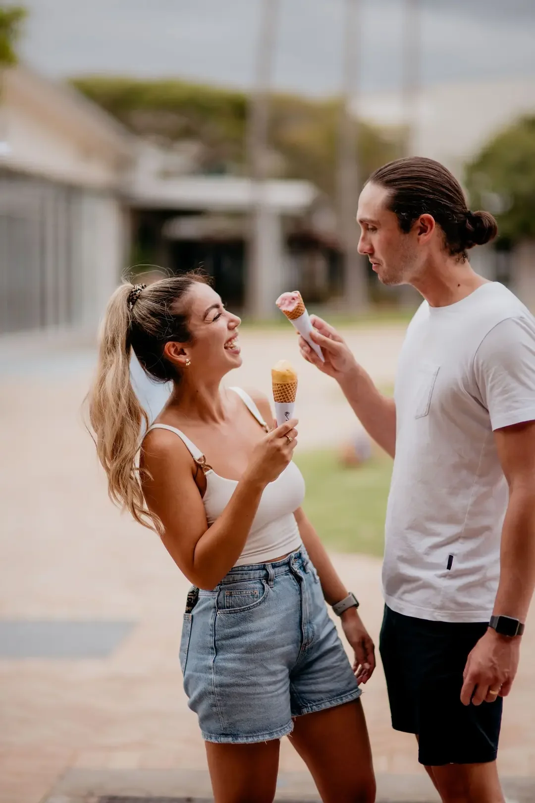 A young woman and man sharing ice cream cones outdoors on a cloudy day, smiling and looking at each other.