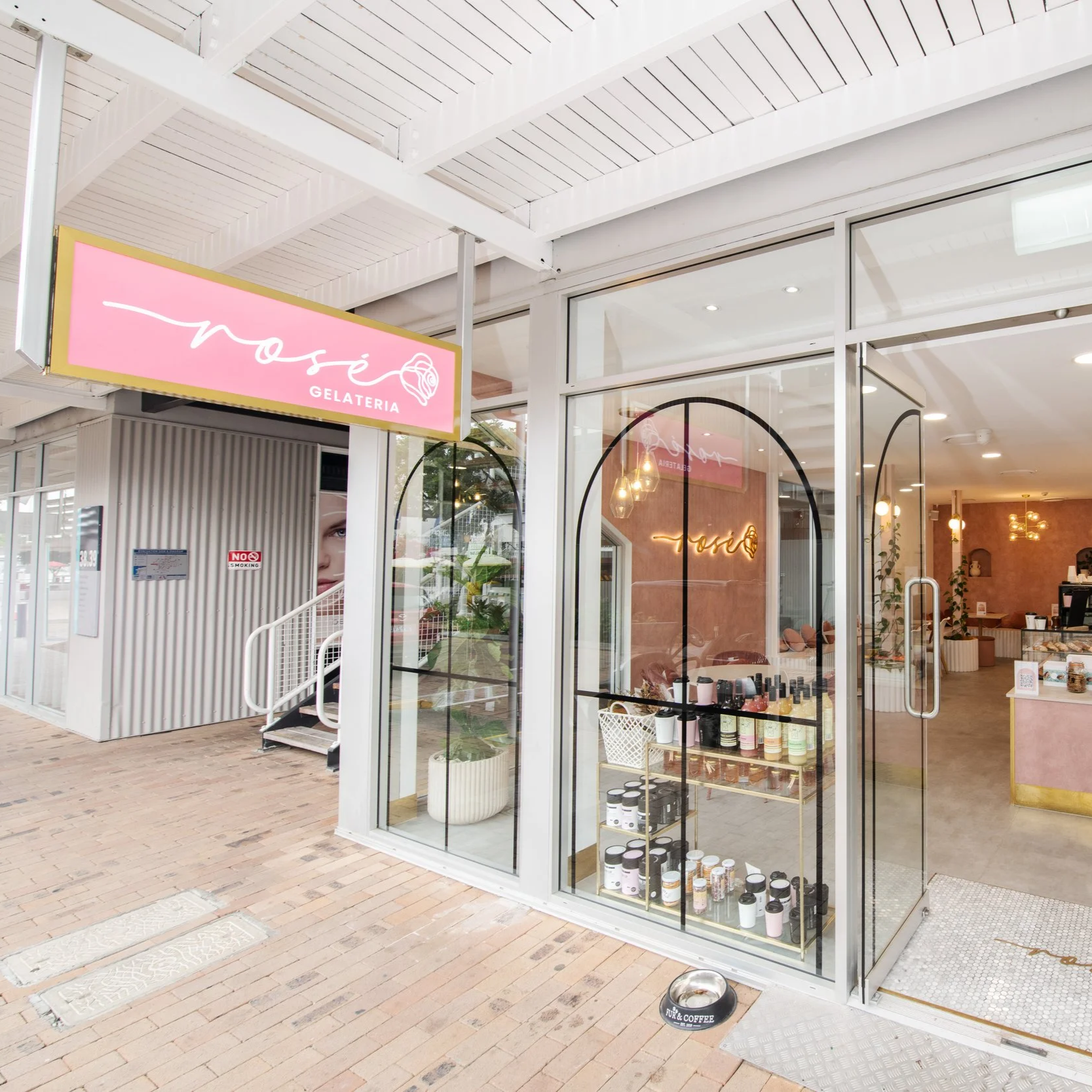 Exterior of a gelato shop named 'Nose Gelateria' with glass doors, displaying shelves with products inside, and a pink sign with white cursive writing and a rose illustration. There is a dog bowl on the sidewalk outside.