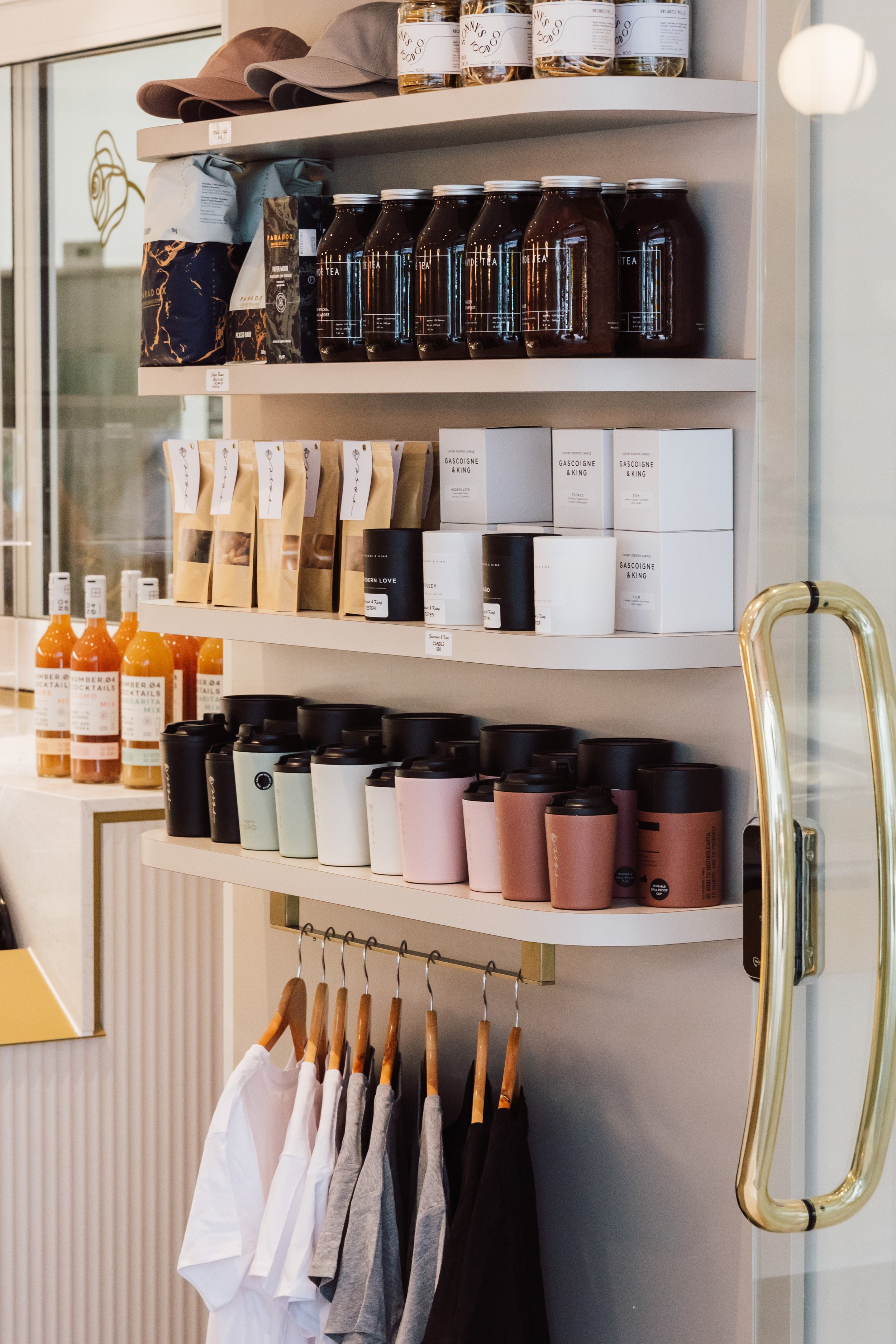 Shelf with hats, jars, coffee and tea containers, and bottles of condiments, with a clothing rack below holding white, gray, and black T-shirts.