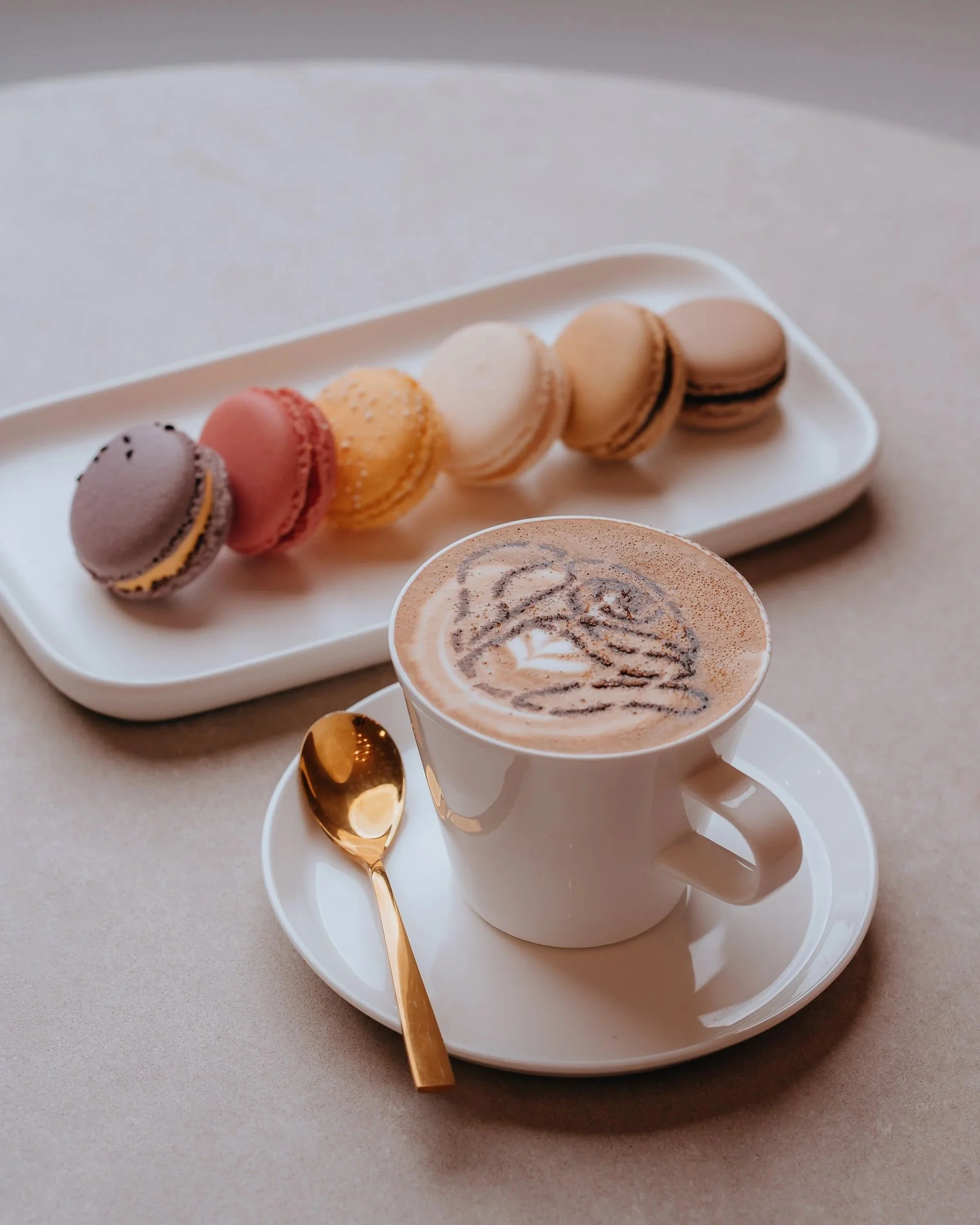 A white cup of frothy cappuccino with a swirl of cocoa powder on top, placed on a white saucer with a golden spoon and a gold-colored stirrer, beside a white plate of colorful macarons in purple, pink, yellow, white, beige, and brown on a light-colored surface.