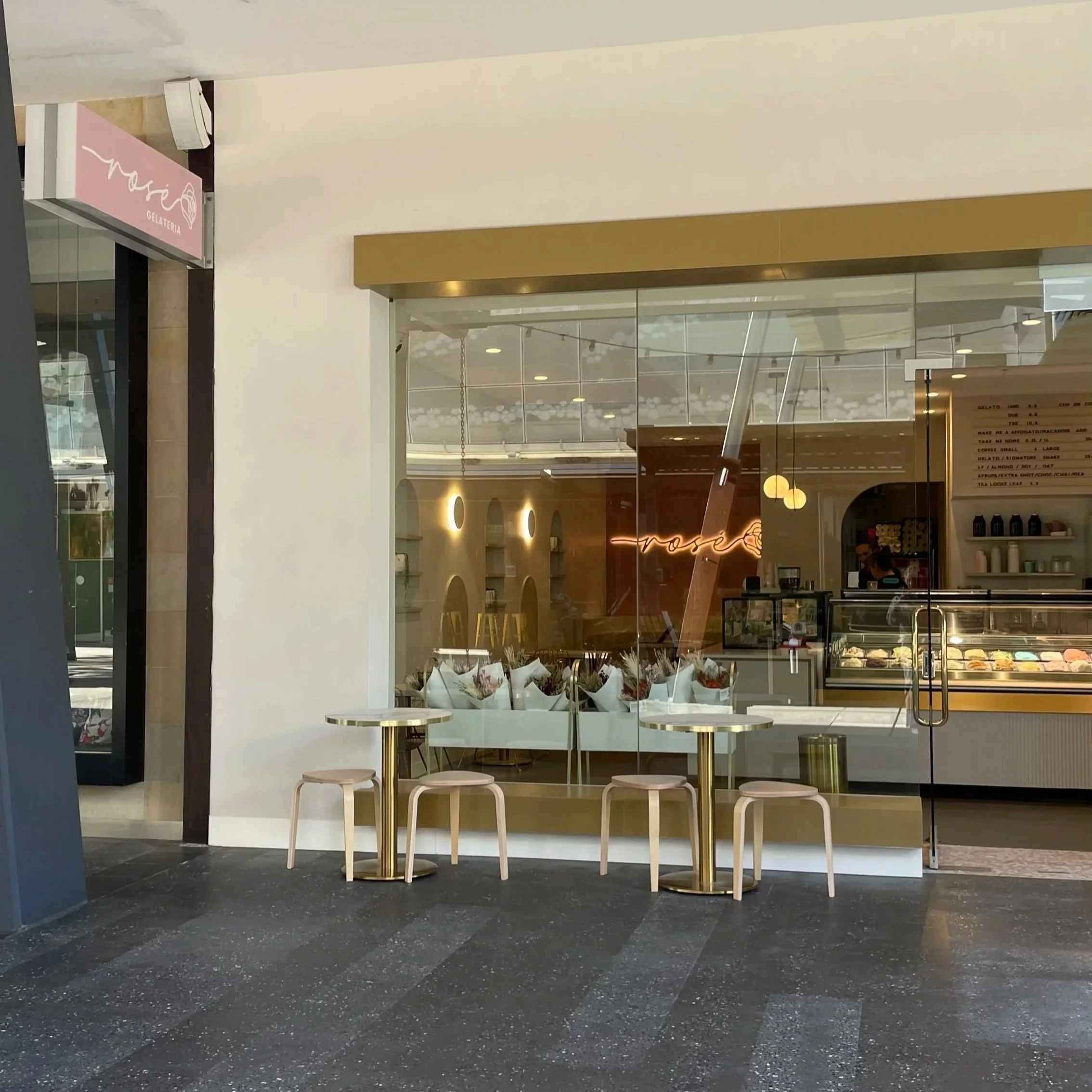 View of a gelato shop with a pink sign and a glass display case showing various flavors of gelato, inside a modern shopping mall.
