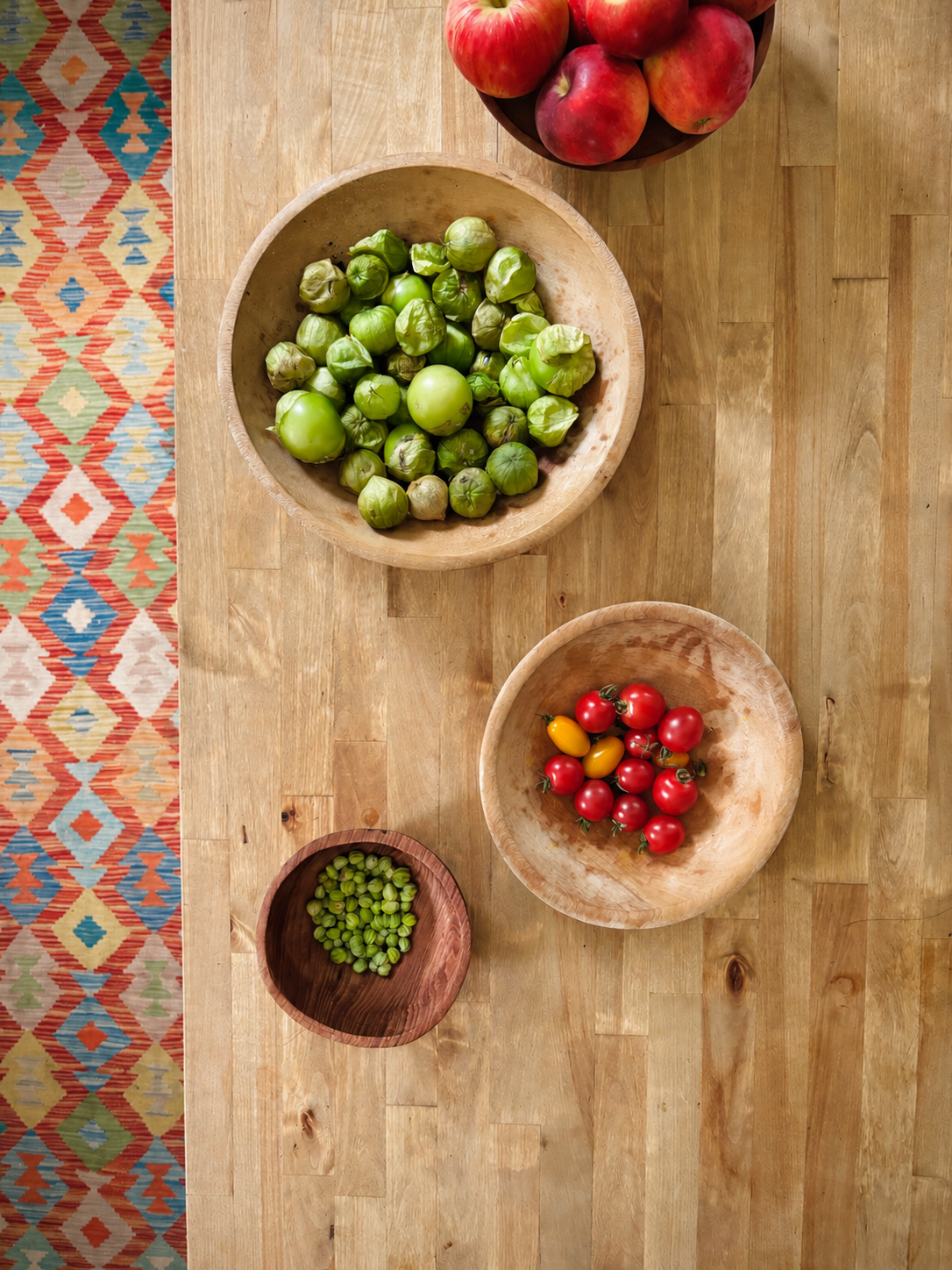 Various fresh vegetables and fruits in wooden bowls on a wooden table, including green tomatoes, cherry tomatoes, and small green peas.