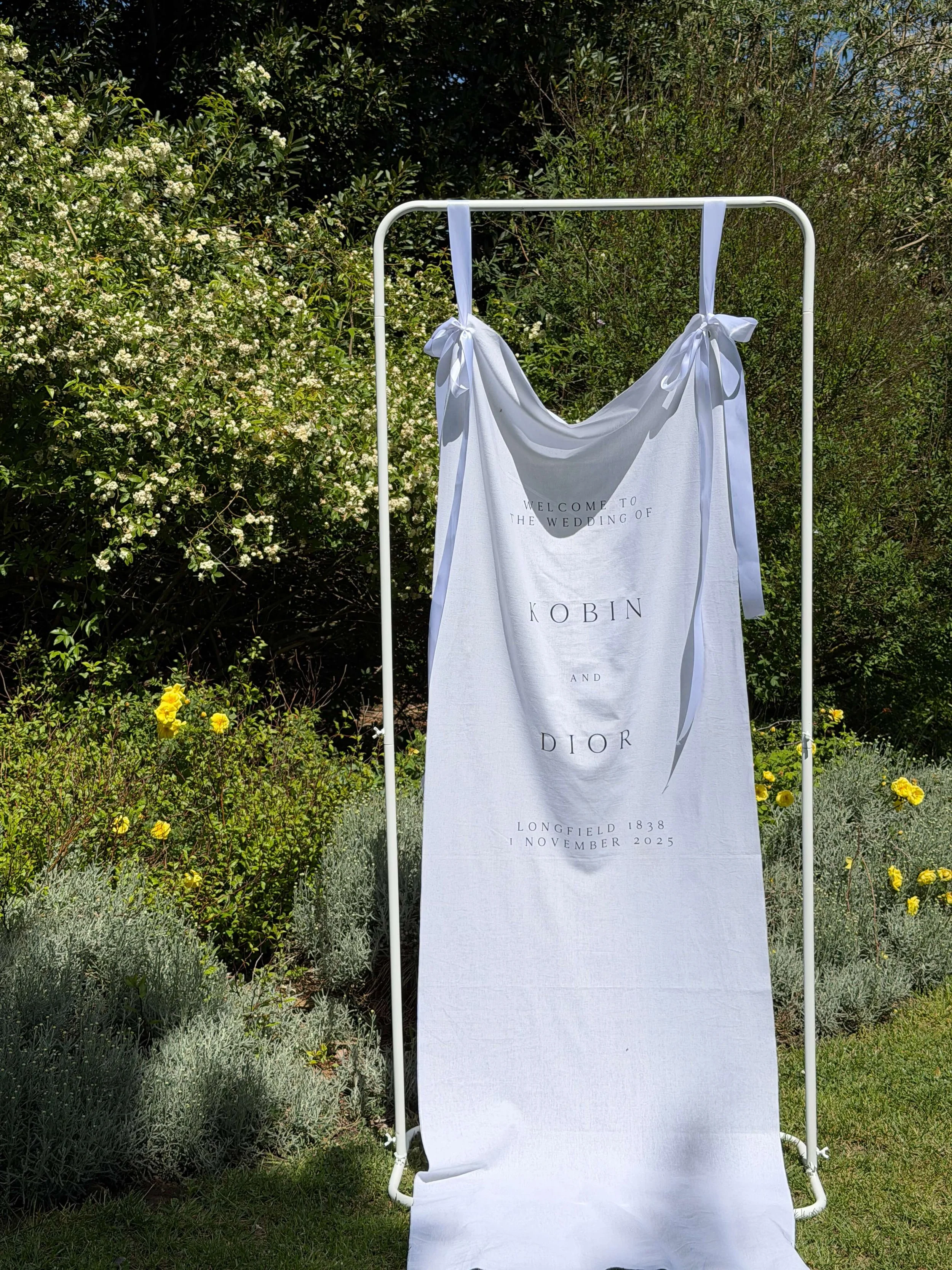 White fabric banner with black text on a white stand, surrounded by green bushes and plants, with yellow flowers in the foreground.