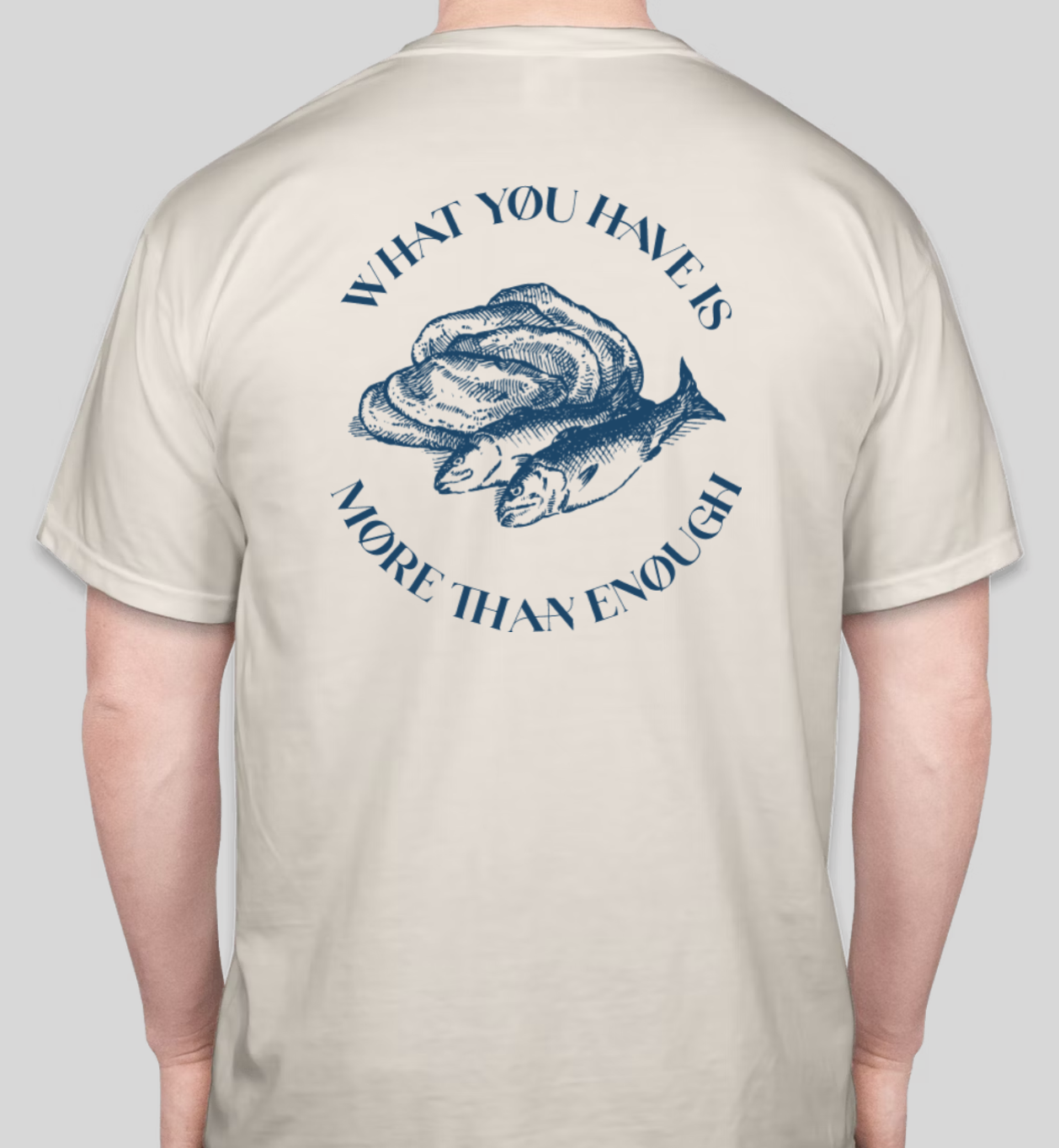 Back of a white T-shirt with a blue illustration of a hand holding a shell and fish. The text reads 'What you have is more than enough' in a circular pattern around the illustration.