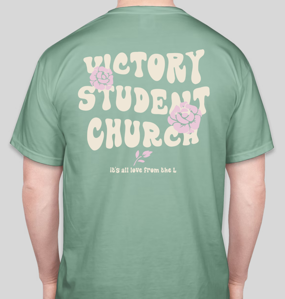 Green t-shirt with cream-colored text that reads 'VICTORY STUDENT CHURCH'. The shirt features pink floral illustrations and smaller text below that says 'it's all love from the L'.